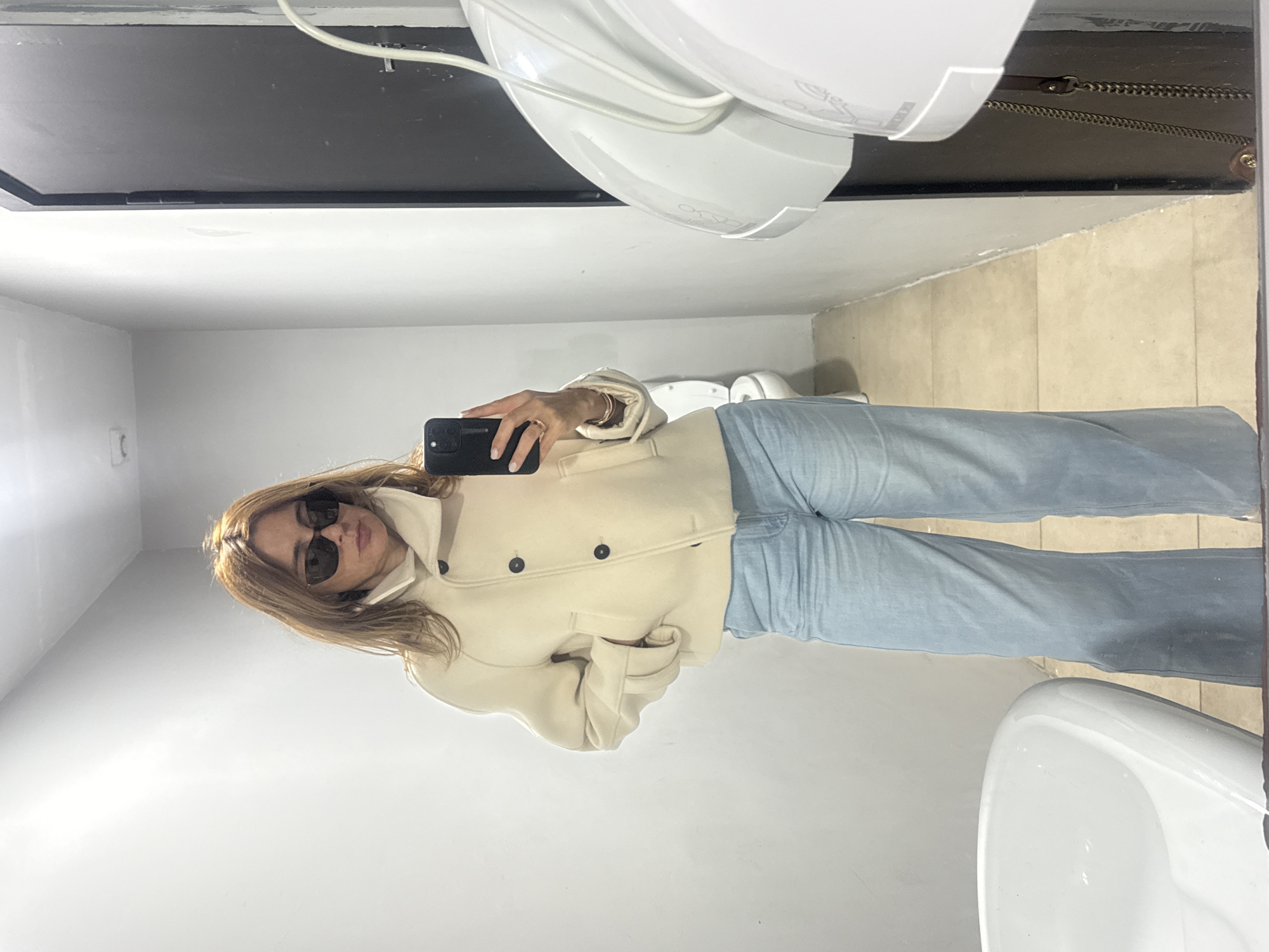 A person stands in a bathroom taking a selfie in a mirror. The individual is wearing blue jeans, a cream-colored coat, and sunglasses. A black smartphone is held in the right hand. The bathroom appears to be a public facility with a white sink, tiled floor, and a black-framed mirror. The image provides no information to determine whether this is in Porto, Portugal.