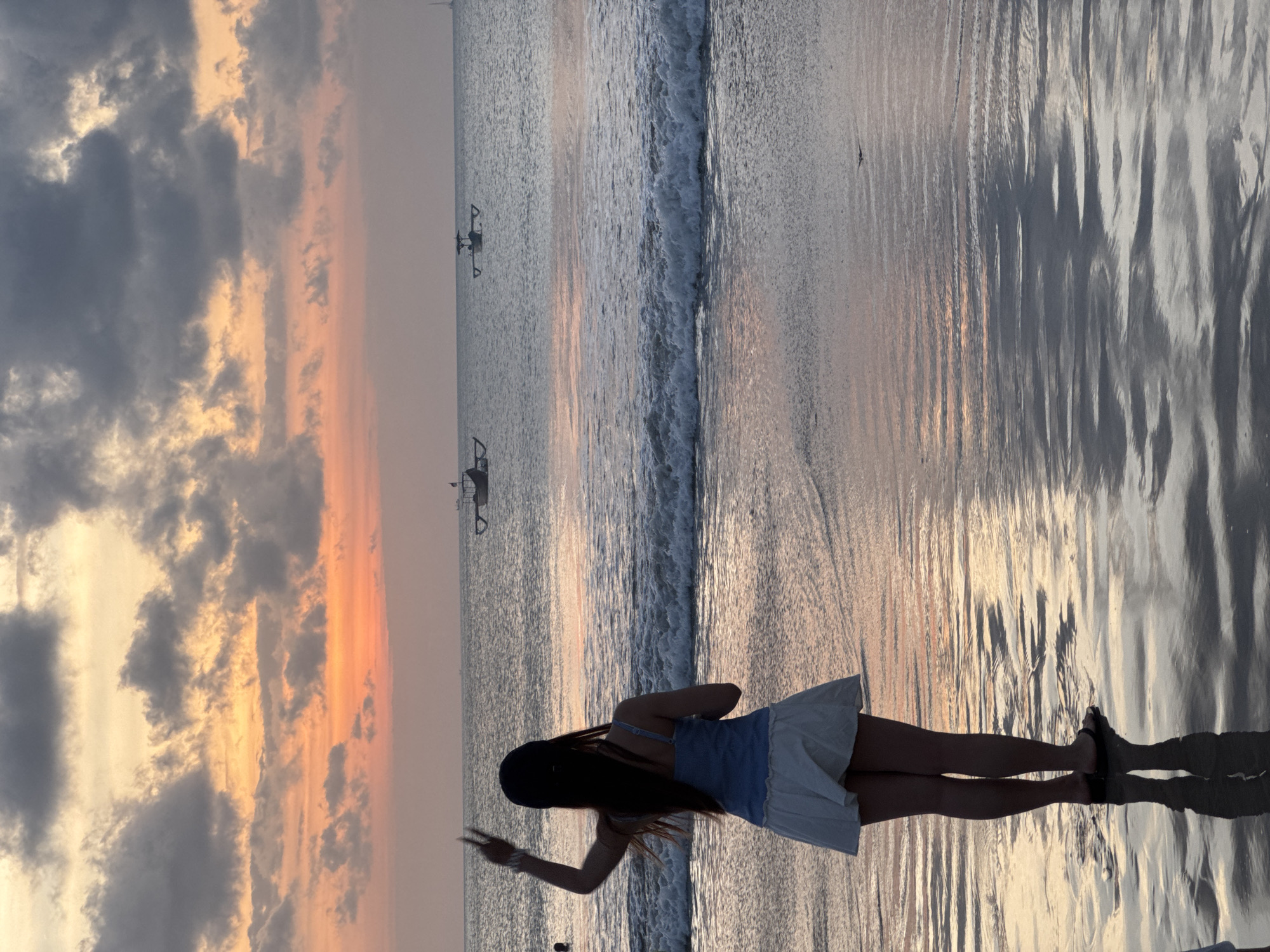 A lone woman stands on a beach in Kuta Selatan, Indonesia, facing the ocean at sunset. The sky is a dramatic canvas of orange and pink hues, with dark, textured clouds streaked across. Gentle waves are breaking on the shore, their white foam reflecting the warm light. The wet sand mirrors the colors of the sky, creating a shimmering, reflective surface. In the distance, two traditional Balinese fishing boats, known as jukung, are visible on the calm sea. The woman, seen from behind, wears a blue tank top and a white skirt, her dark hair flowing down her back. She has a black baseball cap on her head and is making a peace sign with her left hand, seemingly enjoying the serene view.