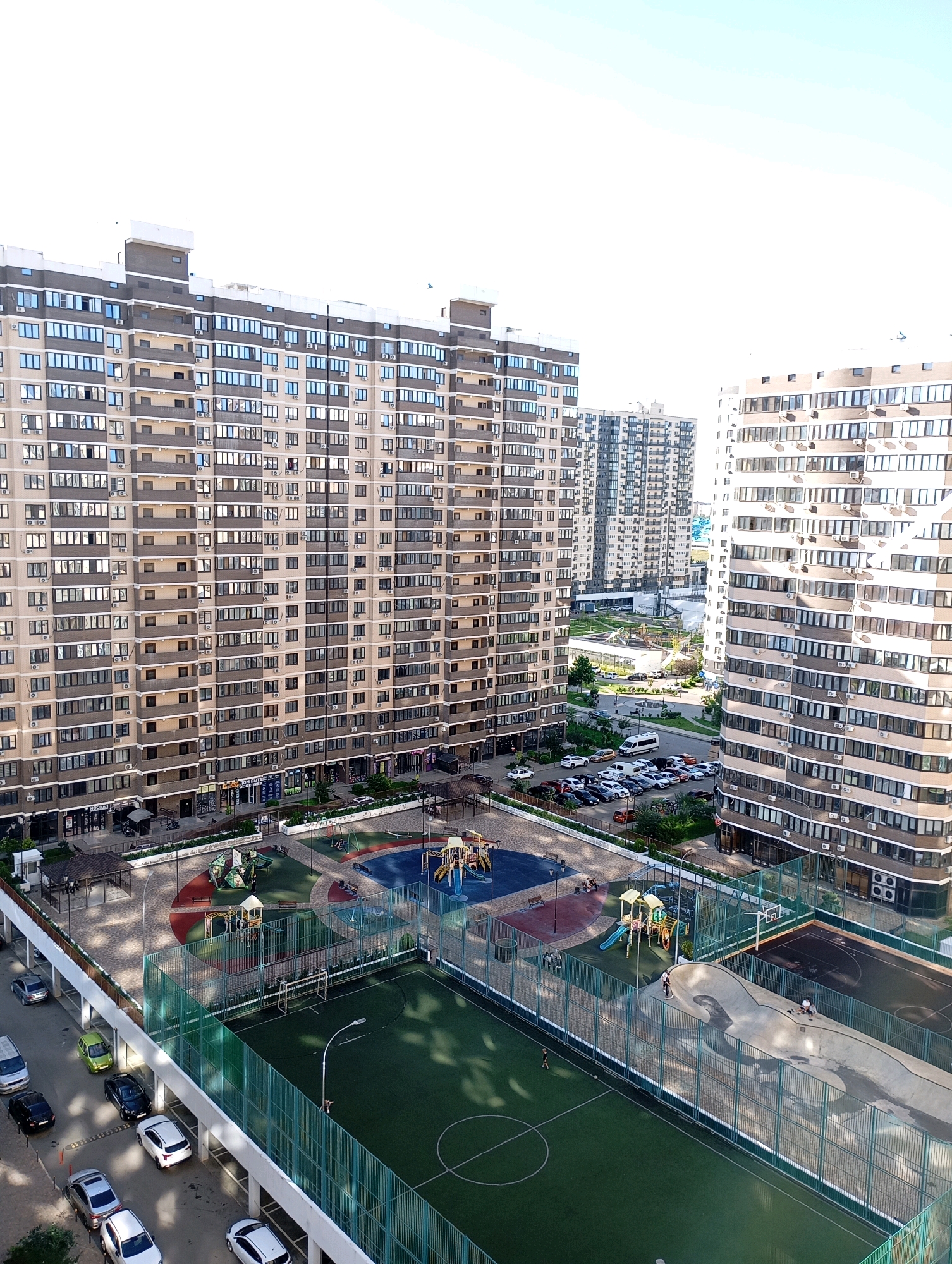 The image depicts a modern residential complex in Krasnodar, Russia. The scene is dominated by tall, multi-story apartment buildings with numerous windows. In the foreground, there is a sports complex featuring a soccer field with artificial turf, a basketball court, and a skate park. Several people are visible in the sports areas, including individuals playing soccer, skateboarding, and sitting. Adjacent to the sports facilities is a children's playground with slides and swings. Parking lots filled with cars are located between the buildings and the sports amenities. The environment appears to be a sunny day with a clear sky.