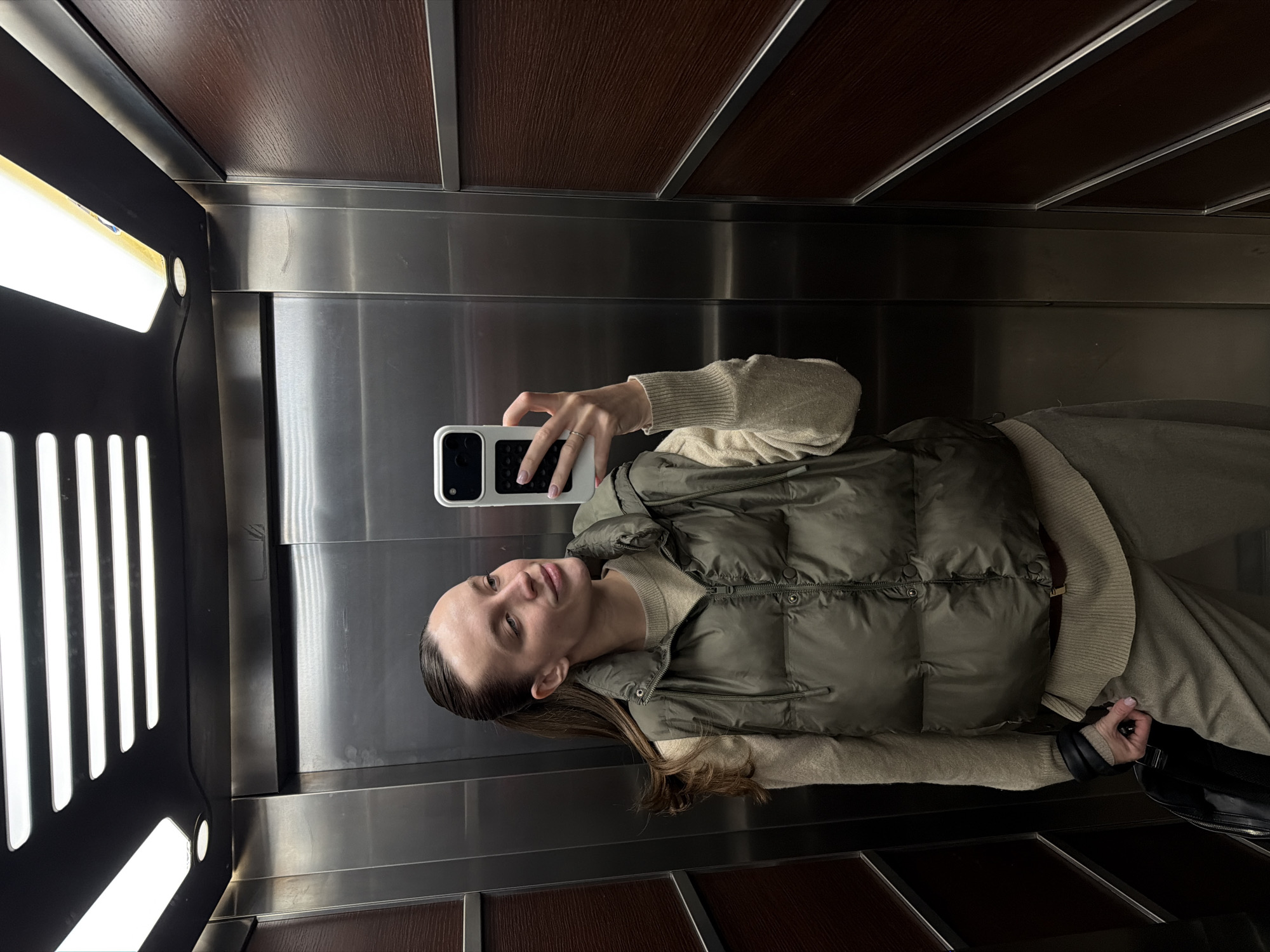 A woman stands in an elevator, taking a selfie with a smartphone. She's wearing a light beige sweater and an olive green padded vest. The elevator interior features metal paneling with wooden trim above and below the central metal sections. The lighting appears to come from a vertical light fixture to the left. The location is likely inside a building, but no specific identifying details indicate it is in Budva, Montenegro.