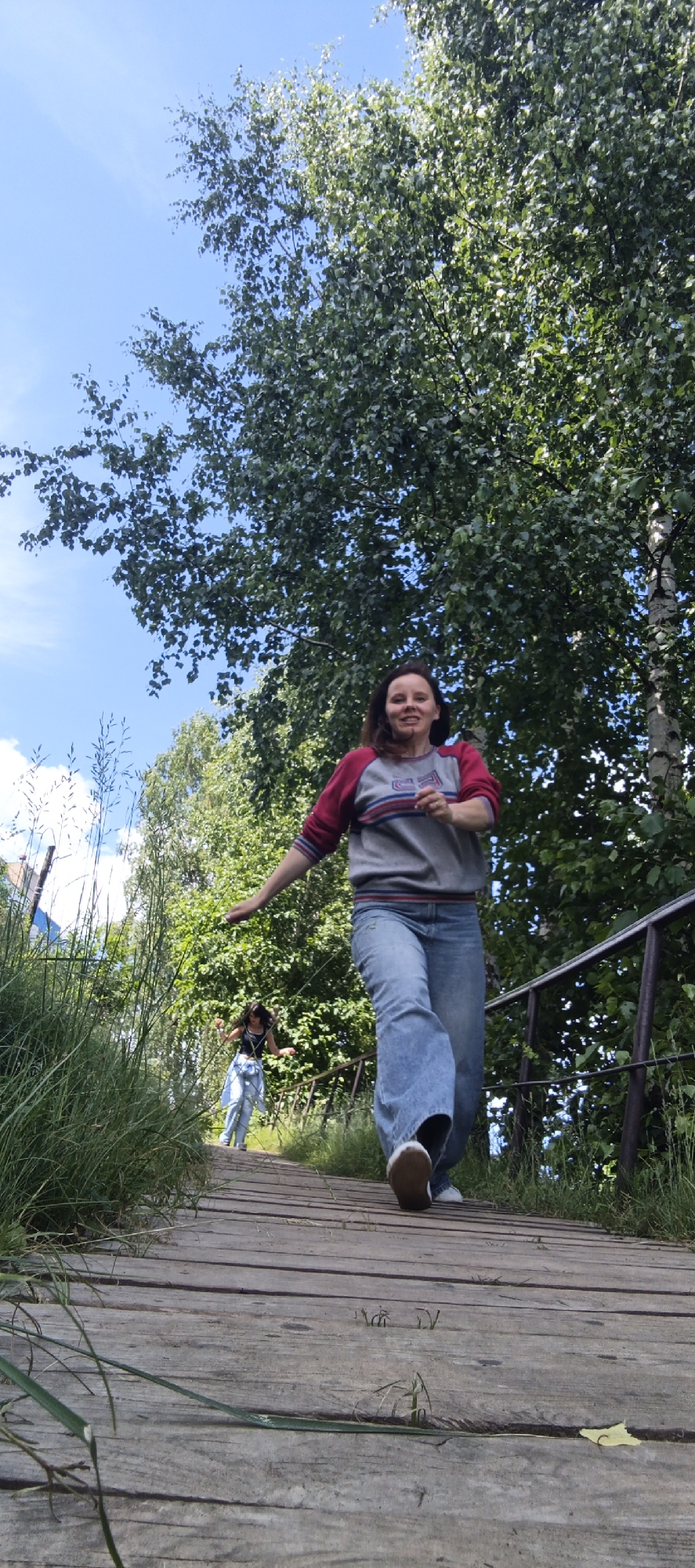 A full-body shot captures a woman in her mid-30s in the foreground, her gaze directed towards the camera with a smile as she walks down a wooden plank pathway. She is dressed in a grey sweatshirt with burgundy sleeves and blue denim jeans. Behind her, further down the path, a second, younger woman, possibly in her late teens, is also walking. She is wearing a black tank top and light blue jeans. The pathway is bordered by tall green grass on the left and a metal railing on the right. The scene is set outdoors, with a large tree with dense green foliage dominating the background against a clear blue sky. The lighting suggests it is daytime, possibly late morning or early afternoon.