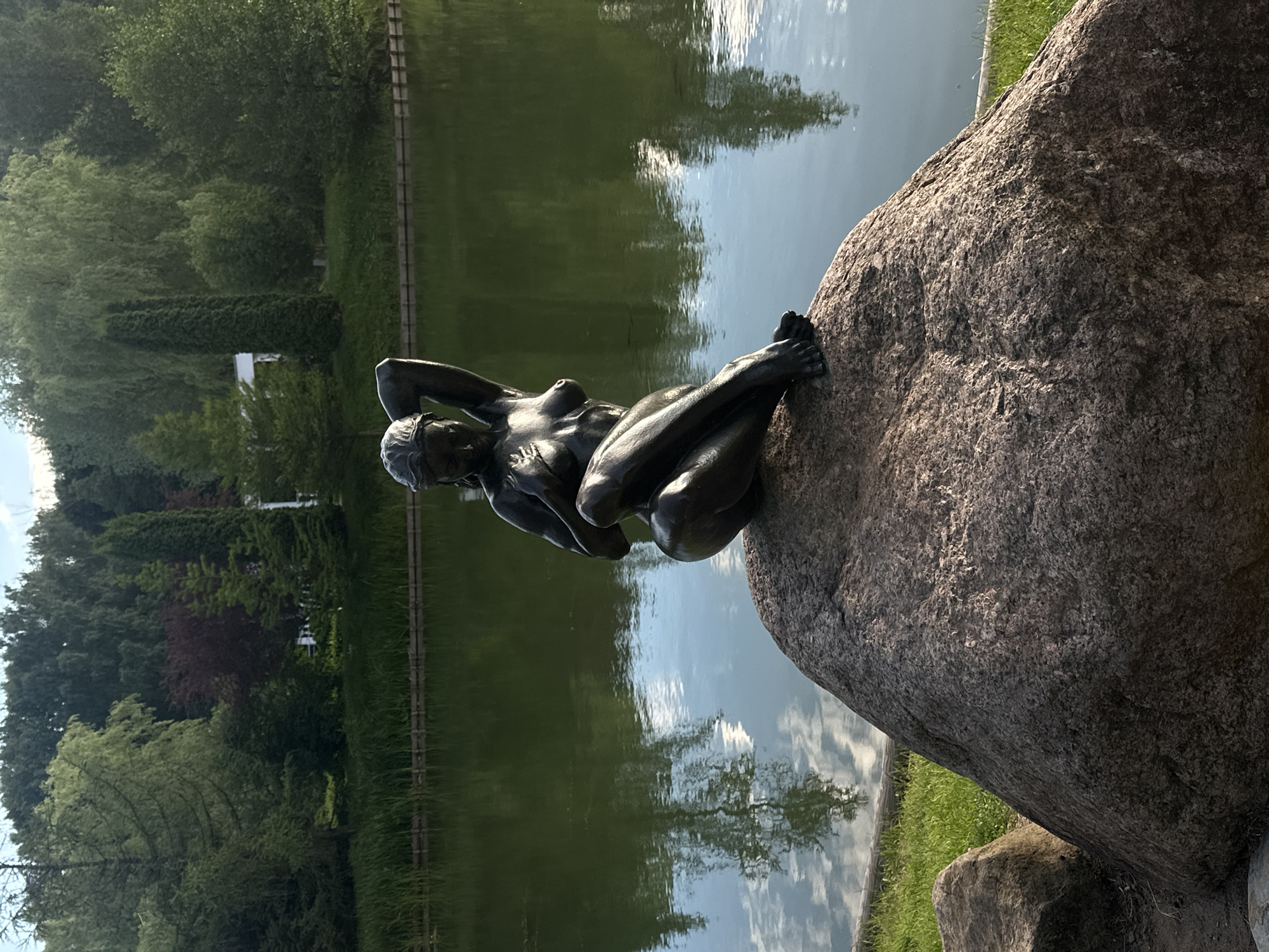 A dark bronze statue of a nude woman is perched on a large, textured boulder. The woman is posed with her left arm behind her head and her right arm resting on her left knee. Her legs are crossed at the ankles. The statue is positioned in front of a calm body of water, which reflects the surrounding trees and the sky. The background consists of a lush green landscape with various trees, including some with dark red foliage. The scene appears to be a park or garden, with the water's edge lined by reeds and a wooden barrier. The lighting suggests it is daytime, with sunlight casting shadows and highlights on the statue and rocks.