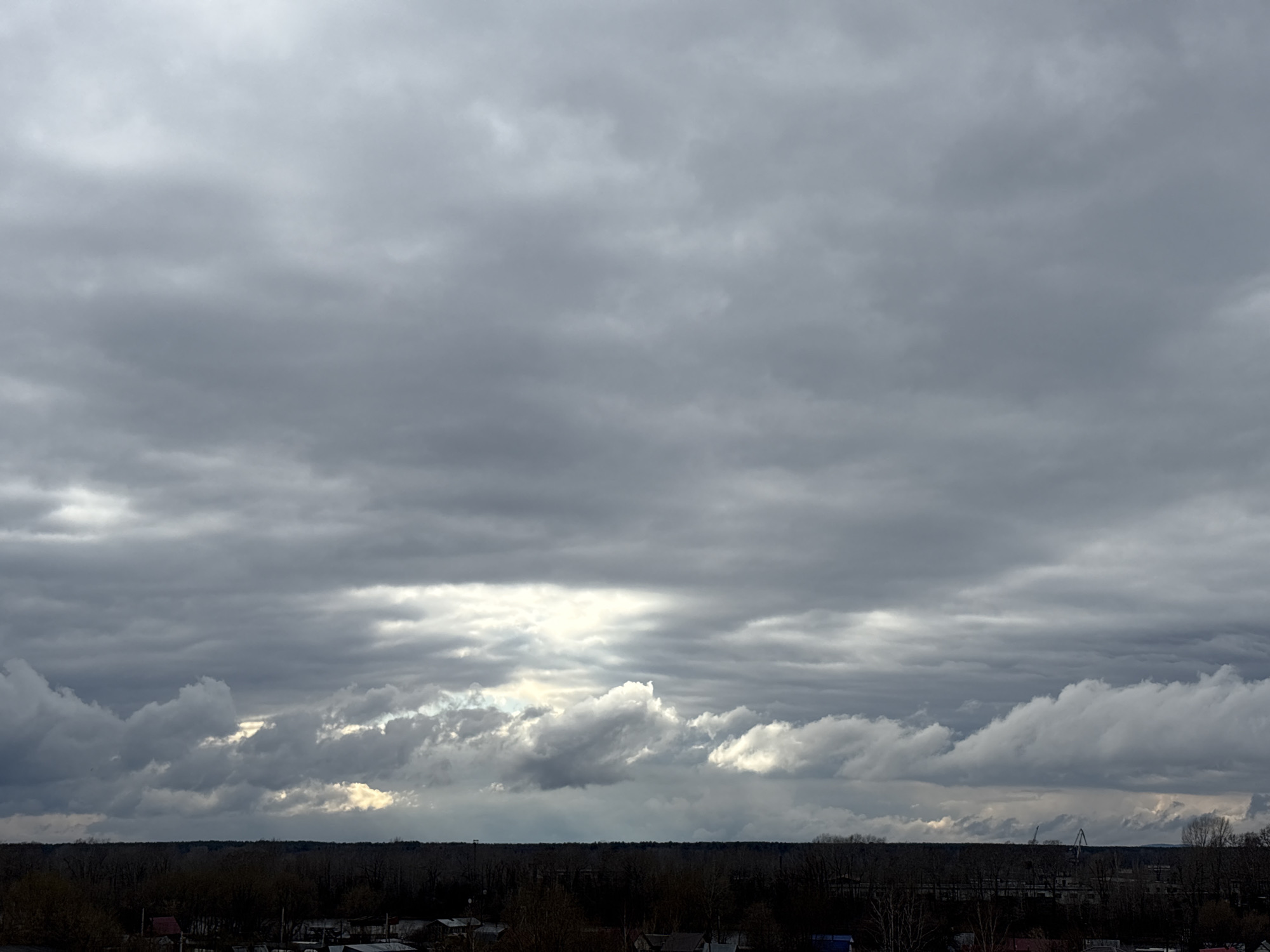 The image presents a wide-angle landscape dominated by a vast, overcast sky, stretching above a distant horizon line. The sky is filled with multiple layers of grey clouds, varying in shade from darker, dense formations in the upper and middle sections to lighter, more broken clouds closer to the horizon. Towards the center and right, brighter, almost white patches of light are visible where sunlight penetrates or peeks through gaps in the lower cloud layers, creating a striking contrast against the general gloom.
A dark, dense band of trees or forest forms a continuous line across the entire horizon, appearing largely silhouetted. Below this tree line, faint shapes suggest a scattered settlement, possibly a village or town. On the far right, near the horizon, some tall, slender structures, possibly cranes or industrial installations, can be glimpsed, hinting at an industrial presence near the natural landscape.
The scene depicts a cloudy daytime, with the weather appearing heavy and possibly indicating rain. There is no discernible human activity. The overall impression is one of a tranquil yet somewhat dramatic natural setting under a heavily clouded sky, with subtle indications of human habitation and industry in the distance, consistent with the Chaikovsky, Russia context. No text is visible in the image.