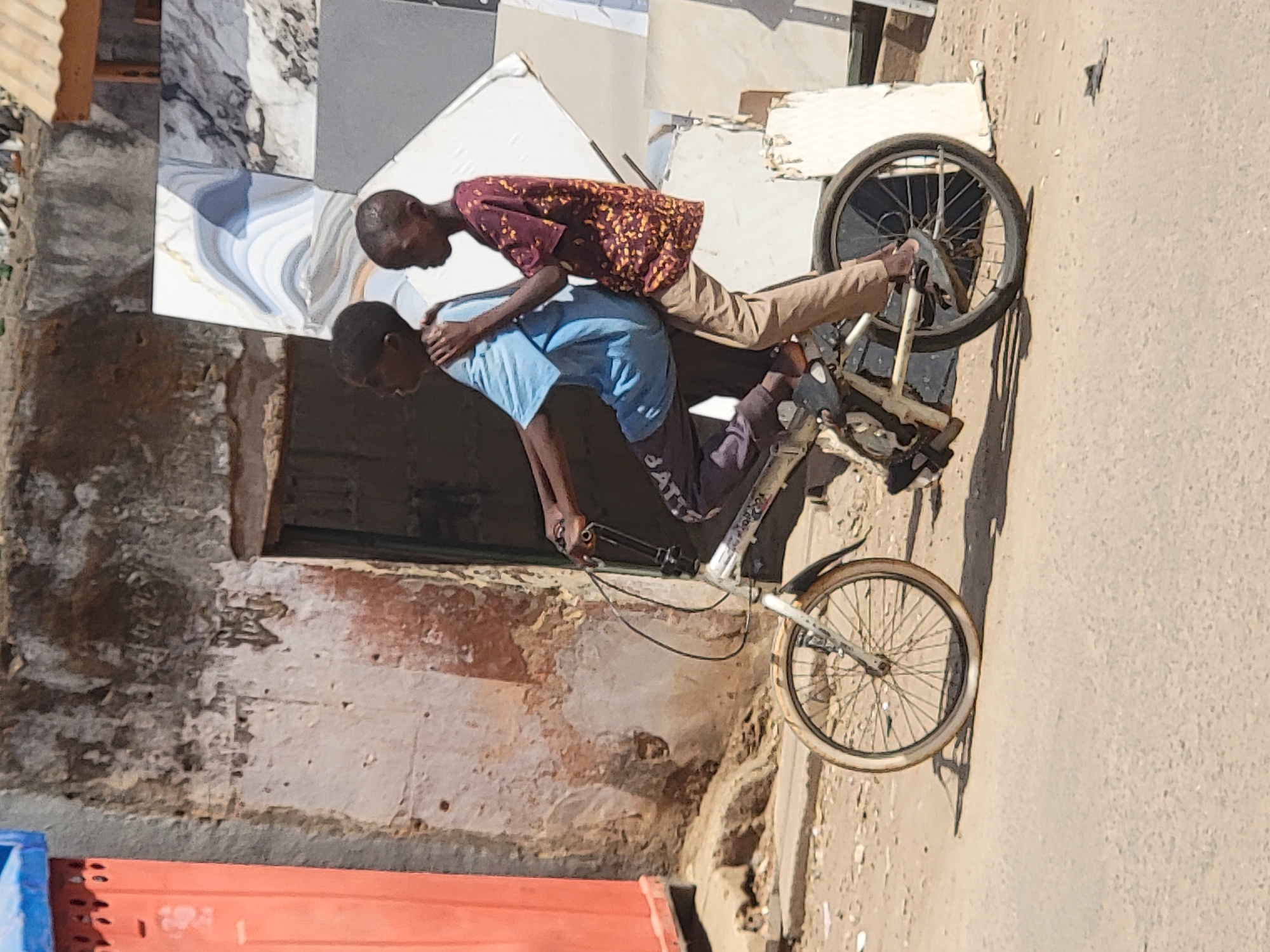 Two young males are precariously riding a bicycle in what appears to be an urban environment. The first male is standing on the pedals and leaning over the handlebars, while the second male is lying across the back of the bicycle, resting his weight on the first male. The bicycle appears to be an older model. The background shows a partially deteriorated building with a mix of concrete and textured surfaces. Tiles lean against the building. The ground is a sandy color. Part of an orange object is visible in the lower left of the frame. The image is taken in daylight. The location is Maiburiji, Nigeria.