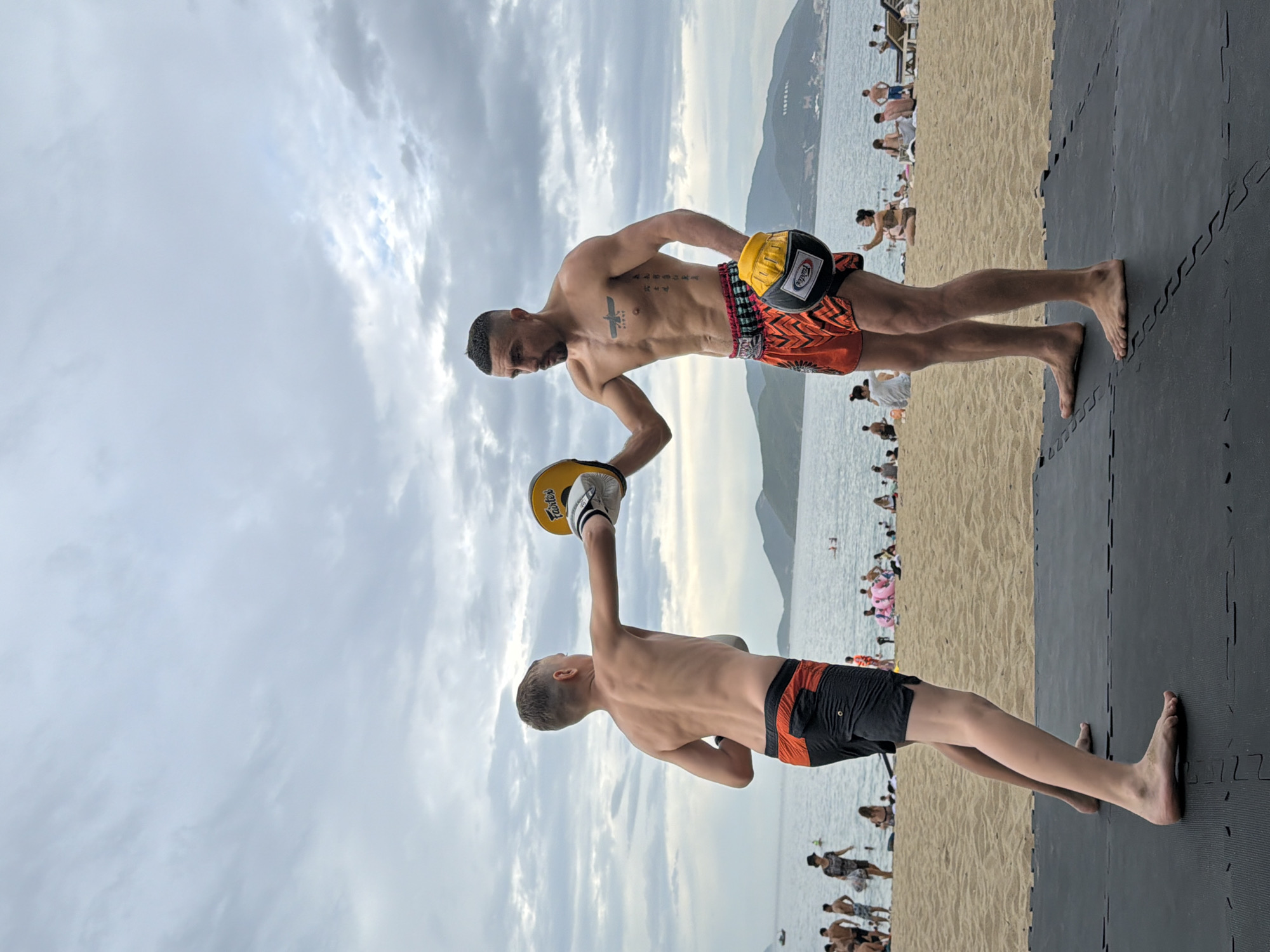 In Nha Trang, Vietnam, two individuals are engaged in a martial arts training session on a beach. A young boy, wearing black and orange Muay Thai shorts, stands on the left, his back to the viewer, throwing a punch with his right fist, which is covered by a white and yellow boxing glove. To his right, a man with visible tattoos on his chest and torso, wearing orange and black Muay Thai shorts, stands facing the boy, holding a yellow punch pad in his left hand, ready to receive the punch. Both are barefoot, standing on a black padded mat on the sand. The background reveals a crowded beach with numerous people relaxing and swimming in the sea, under a cloudy sky. The coastline is lined with hills in the distance.
