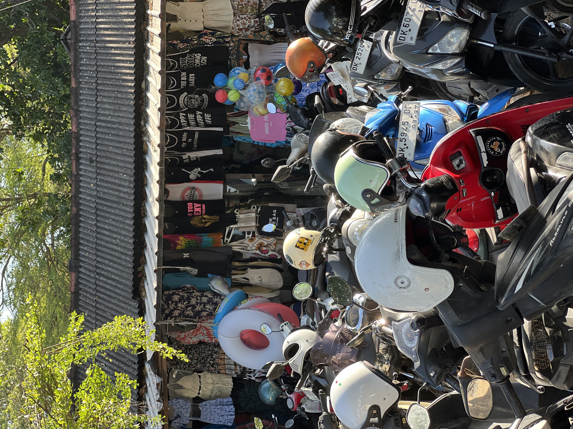 This is a vertical, medium-angle, outdoor shot of a roadside stall in Denpasar, Indonesia, likely taken in the morning or afternoon given the bright sunlight and sharp shadows. The scene is cluttered with a variety of items, primarily motorcycles and clothing. In the foreground, several motorcycles are parked, some with helmets stacked on them. The motorcycles are of various colors, including blue, red, and black. Helmets are a prominent object, with at least four visible, in white, light green, and black. One white helmet has the word "HONDA" on it.
Behind the motorcycles, the stall displays a dense collection of clothing, mostly t-shirts, folded and hanging. Many of the t-shirts feature text and graphics. Visible text includes "TOU TANG EXY THIS SHIRT," "My Wife is Awesome WIFE HAPPY FE," "THE BUM SIX BABYS PACK COMING SON," "WHITE FOX VOL3," "DK 25200 12," "DK 6074," and "K.5945 OP 09-27." There are also some colorful inflatable rings, one red and white and another blue, placed among the clothing. The background shows a corrugated metal roof, partially covered by green foliage from a nearby tree. The overall impression is of a bustling marketplace with a mix of goods for sale, typical of a tourist or local shopping area in Southeast Asia. The sunlight suggests a warm, possibly humid day. No people are clearly visible in the frame.