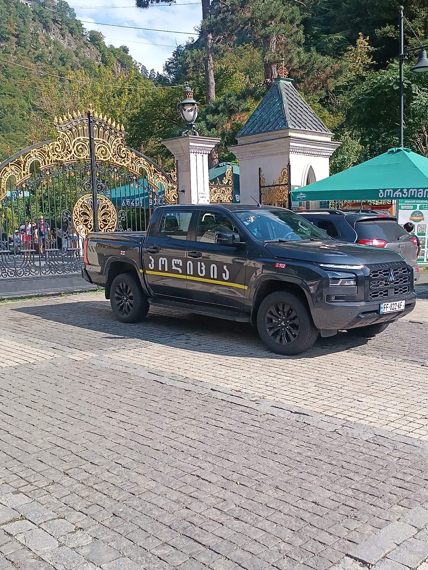 A dark-colored pickup truck with yellow lettering is parked on a cobblestone street in Borjomi, Georgia. The truck has Georgian license plates and appears to be some kind of official vehicle. Behind the truck is a large, ornate golden gate, suggesting the location is a park or a place of significance. Further in the background, there are trees and a hillside, typical of the Borjomi area. A green awning with Georgian lettering is visible to the right, possibly indicating a vendor or establishment. The sky is clear and blue, suggesting a pleasant day.