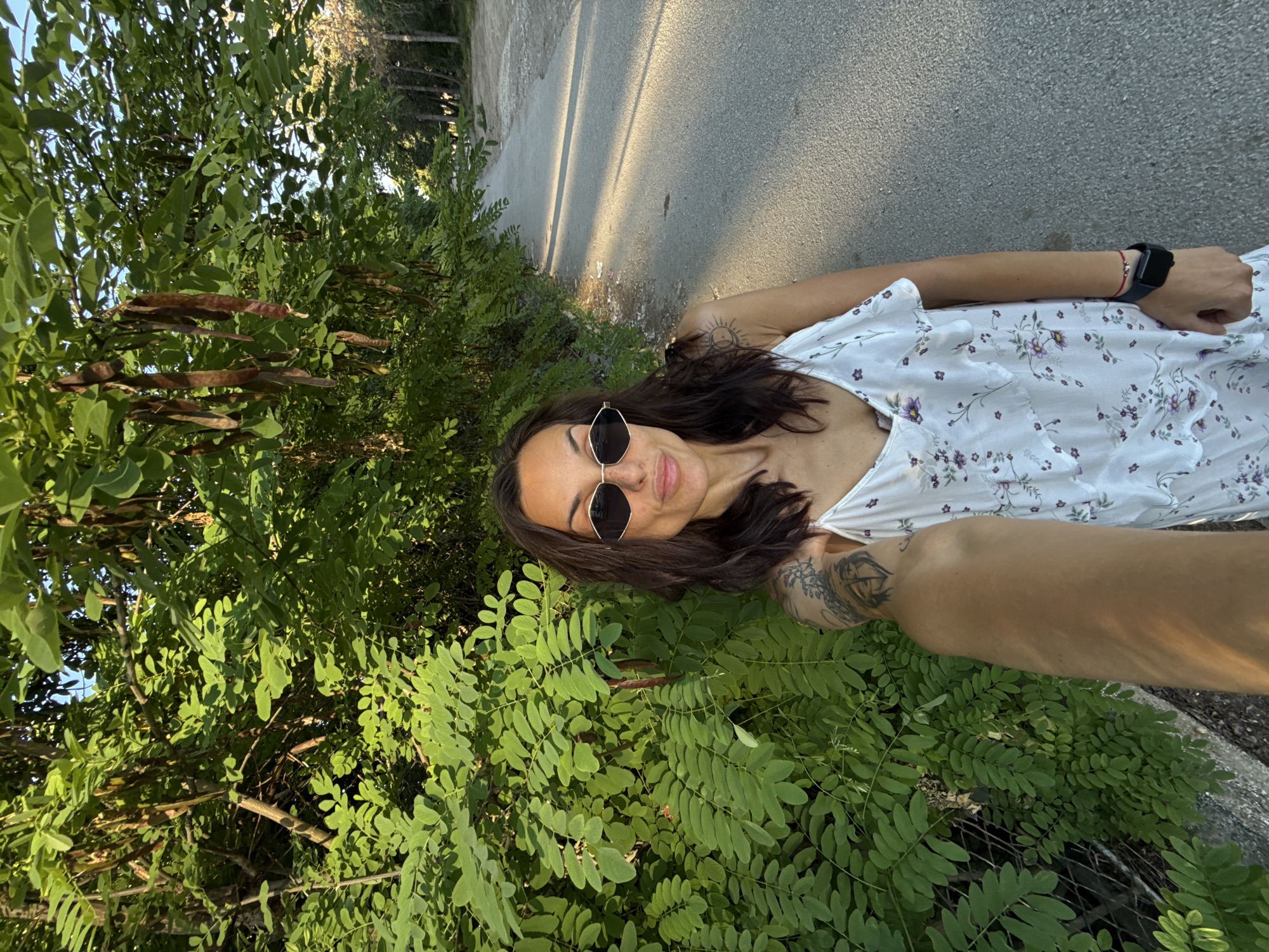 A woman stands outdoors on a paved path, with lush green foliage and trees to her left and the path extending into the distance on her right. She is wearing a white dress with a floral pattern, sunglasses, and a smartwatch. The scene appears to be a park or a tree-lined street in Pleven, Bulgaria, likely during daylight hours due to the sunlit path and the green leaves. The woman is holding up her arm as if taking a selfie.