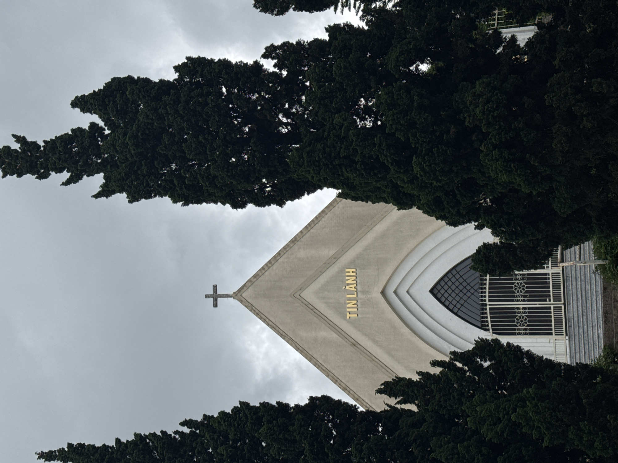 A low-angle, vertical shot captures a church partially obscured by dark green trees, with a grey, overcast sky in the background. The church has a pointed gable roof, with a cross at the apex. The building's facade is primarily a light gray color, featuring white architectural detailing around a large window that is barred with intricate metalwork. Vertically oriented, gold letters reading "TIN LANH" are visible on the facade. The dense foliage of the dark green trees frames the structure on both sides, partially concealing the church and adding depth to the image. The architecture appears modern and minimalist in design, while the trees give the image a naturalistic element. This building is a Protestant Church located in Da Lat, Vietnam.