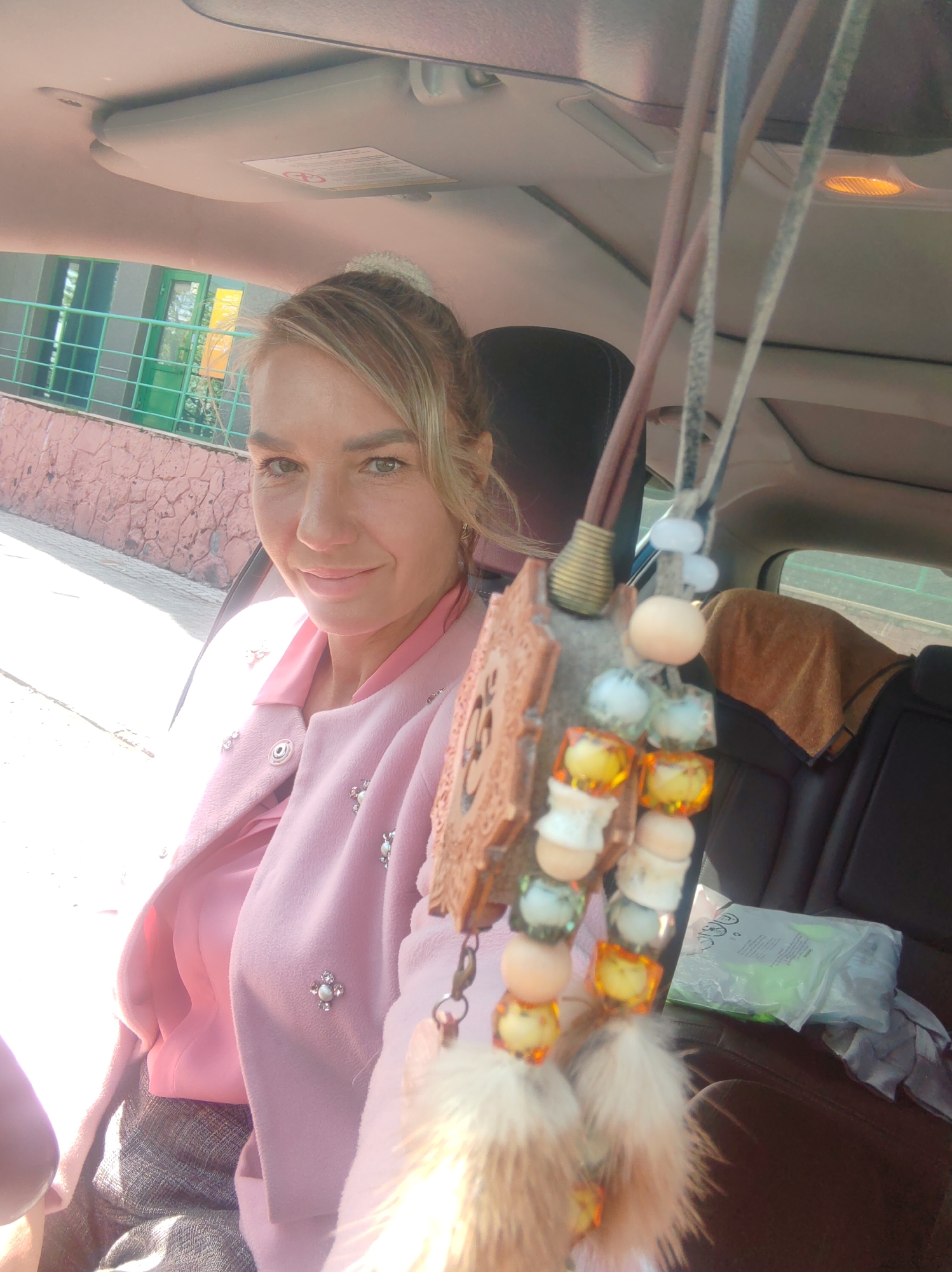 A medium shot captures a woman sitting in the passenger seat of a car. She is looking towards the camera with a slight smile. She has blonde hair pulled up in a bun and is wearing a pink collared shirt and a light pink jacket adorned with pearl-like embellishments. In the foreground, a car accessory hangs from the rearview mirror, featuring beads and fluffy charms. The background shows the interior of the car, including seats, and through the windows, an exterior scene with a building and greenery is visible. The lighting suggests it is daytime.
