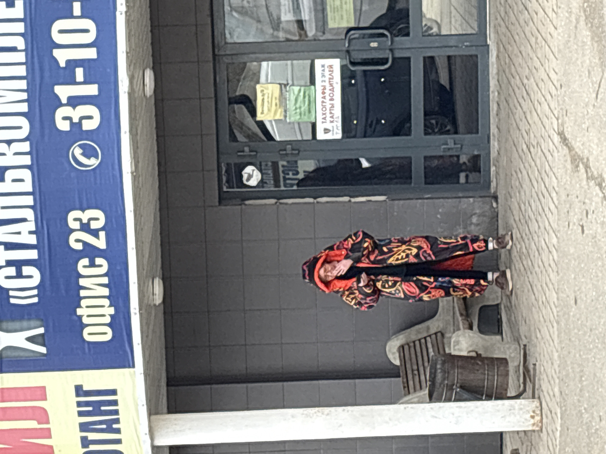 The image depicts an outdoor urban scene in front of a commercial building, located in Izhevsk, Russia. A person stands centrally on a dark gray tiled ground, slightly facing away from the camera. They are wearing a distinctive, brightly patterned hooded coat in shades of red, orange, yellow, and black, with their face partially obscured by the hood and a hand raised.
The building entrance features glass double doors with dark frames, to which several signs are taped. A prominent white sign clearly reads in Cyrillic: "ТАХОГРАФЫ 2 ЭТАЖ" (TACHOGRAPHS 2nd FLOOR), "КАРТЫ ВОДИТЕЛЕЙ" (DRIVER CARDS), and "Тесла" (Tesla), indicating services related to vehicle tachographs and driver cards. Other smaller, less legible yellow and green signs, along with a sticker featuring a musical note, are also on the door. The building's facade around the entrance is a dark gray tile, transitioning to a lighter, textured brick wall on the right side of the frame.
To the right of the person, a wooden slatted bench and a dark wooden barrel or planter are visible. Overhead, a large blue banner with white Cyrillic text is partially visible, stating: "«СТАЛЬКОМПЛЕ" (likely part of "СТАЛЬКОМПЛЕКТ"), "офис 23" (office 23), and a partial phone number "31-10-". A yellow banner with red text "ТАНГ" is visible below the blue one.
The lighting is diffused, suggesting an overcast or cloudy day, with no harsh shadows. The scene portrays a typical daytime commercial street setting.