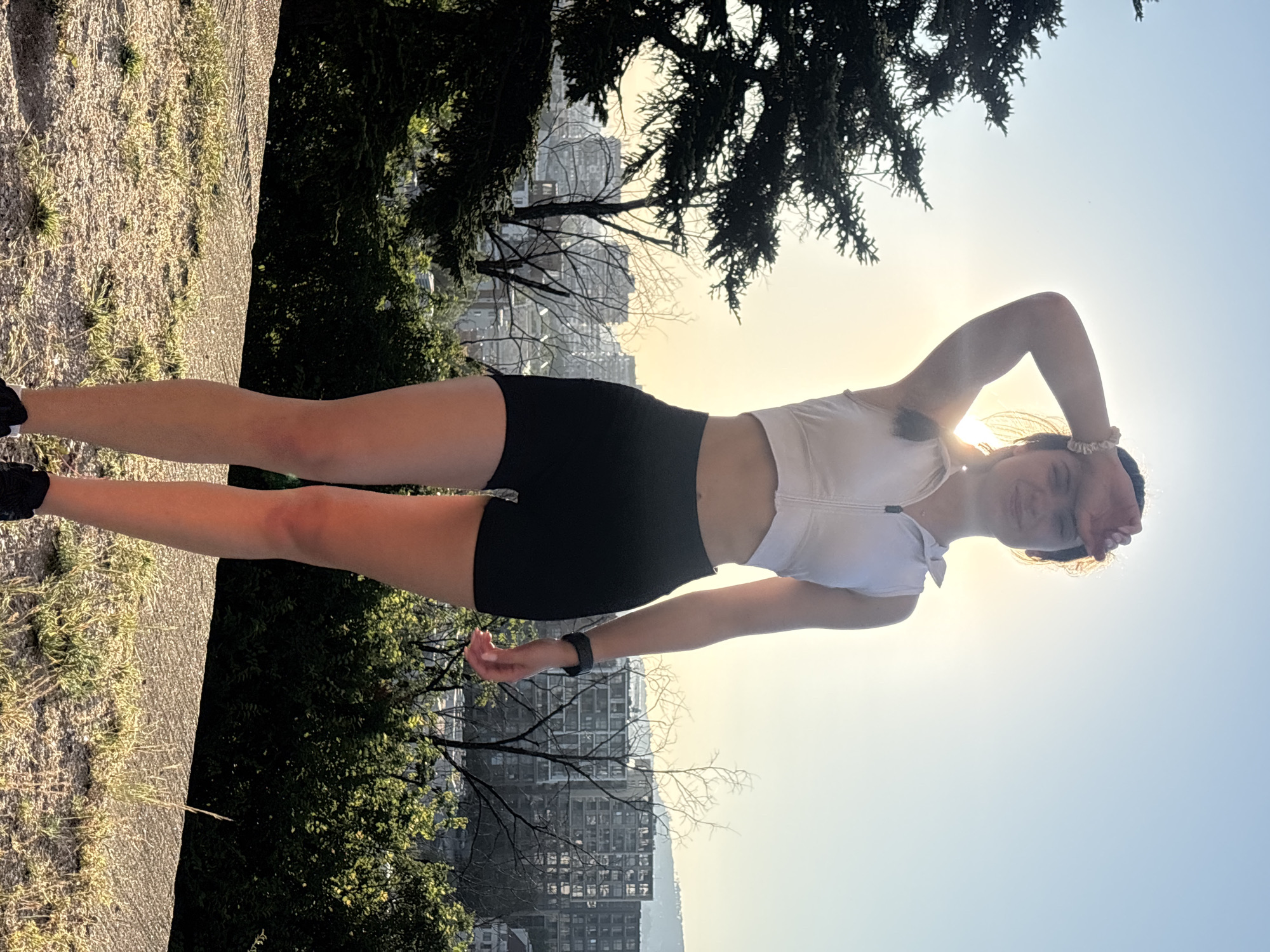 A young woman is captured in a vertical, full-body photograph. She is standing outdoors, in what appears to be a park or elevated area, with a cityscape visible in the background. The sun is setting or rising, casting a warm, golden light and creating a silhouette effect. She is wearing athletic attire: a white, cropped zip-up top and black shorts. Her hair is pulled back, and she has a fitness tracker on her left wrist. Her right arm is raised and resting on her head, with her left hand casually by her side. She is smiling and looking towards the camera, conveying a sense of happiness and relaxation. The background shows trees and the upper sections of buildings, suggesting an urban environment adjacent to a natural space. The sky is clear, indicating fair weather. The overall mood is peaceful and serene, enhanced by the soft lighting of the golden hour. The location is identified as Varna, Bulgaria. There is no visible text in the image.