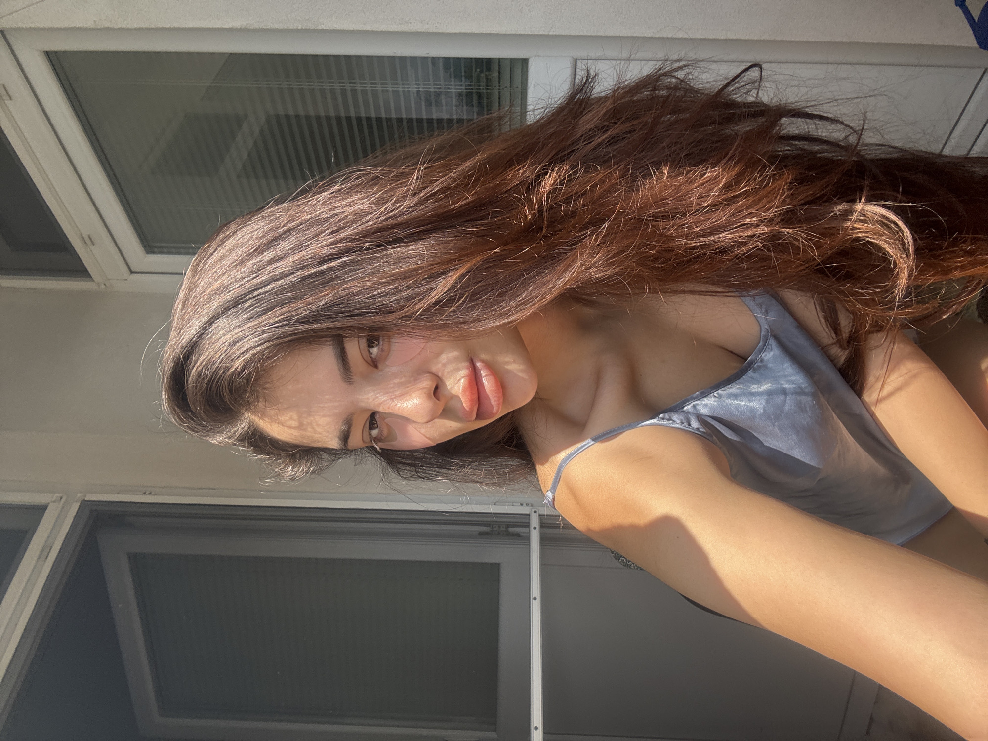 A candid, brightly lit portrait features a young woman with long, flowing dark brown hair. She is captured in a relaxed pose, with her head tilted and a subtle, content smile on her lips, her gaze directed towards the camera. Strong sunlight highlights her features and hair, creating warm, reddish-brown tones and casting distinct shadows on her skin, suggesting it's daytime, possibly morning or late afternoon. She is wearing a light blue or grey silky camisole, visible on her torso and shoulder.
The background shows white-framed windows or glass doors, with reflections hinting at an interior or opposite building structure. The overall impression is one of natural light and an intimate moment. No text is visible in the image. The setting is indoors, with the harsh sunlight indicating a clear day in Plovdiv, Bulgaria.