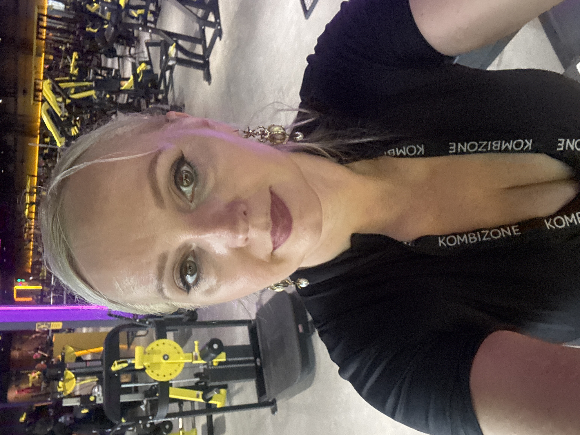 A selfie is taken from the chest up, showing a blonde woman with dark eye makeup and dark lipstick posing in a gym. She is wearing a black zip-up top with "KOMBIZONE" written vertically on it. The background is blurred but shows gym equipment, primarily yellow and black machines. The lighting is dim with some purple and yellow accents. The scene is indoors, consistent with a fitness center. The image does not contain any information to suggest a location in Komsomolsk-on-Amur, Russia.