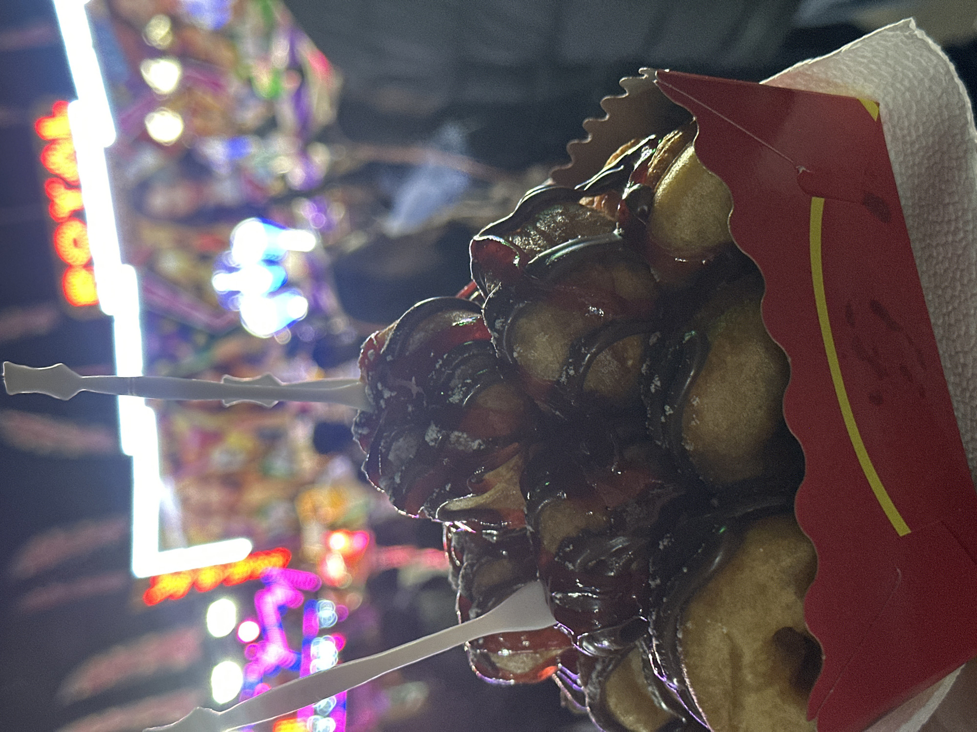 At night, a close-up captures lokma, fried dough balls drizzled with chocolate sauce, skewered by two plastic forks and held in a red and yellow cardboard container with a white paper liner. In the blurred background, lights from an amusement park ride glow in colors like red, white, and blue. The image appears to have been taken at an amusement park. The specific location in the city of Lovech, Bulgaria cannot be determined without additional information.