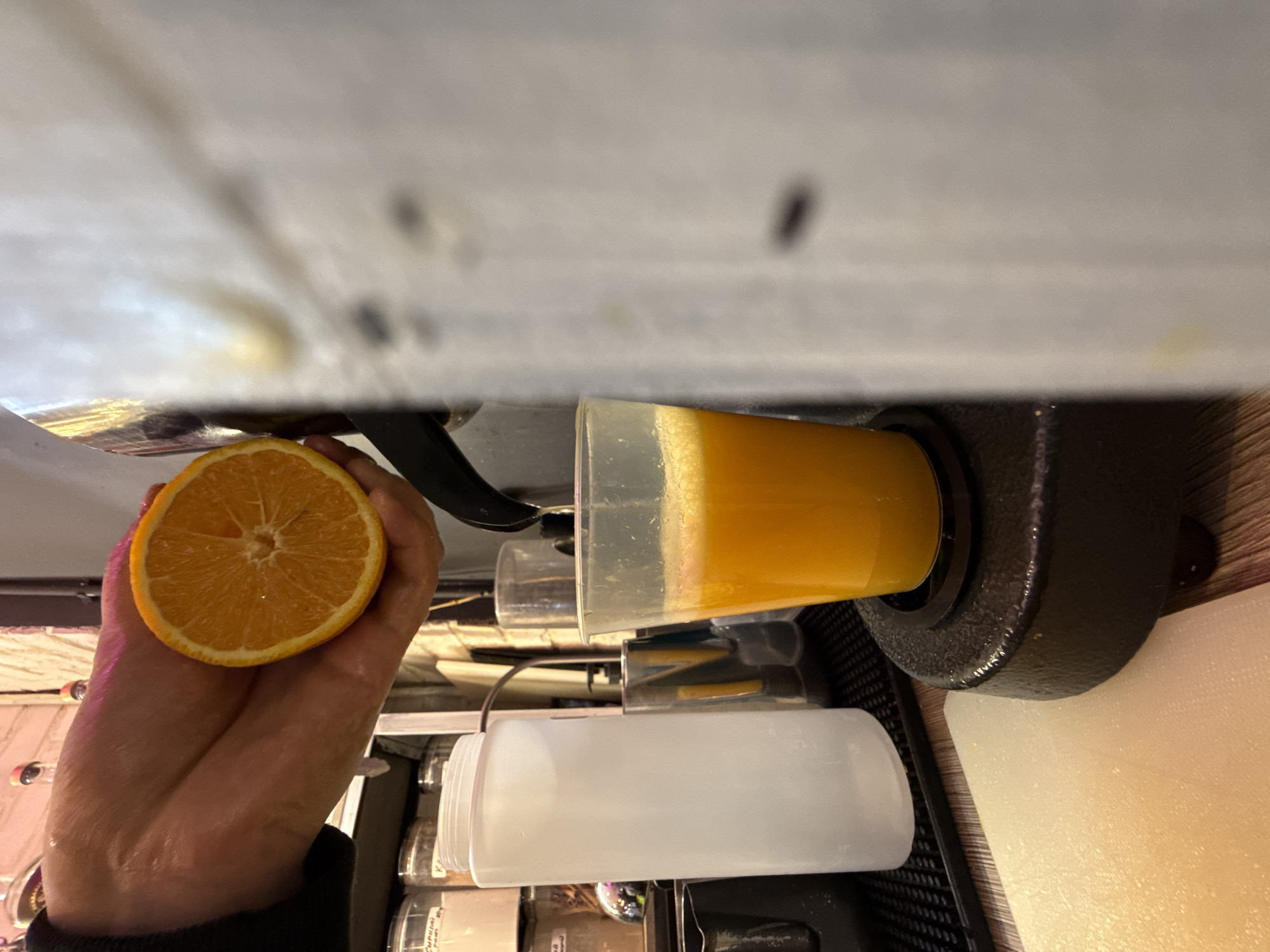 In Moscow, Russia, a hand is holding a halved orange over a clear plastic cup filled with freshly squeezed orange juice. The juice is being dispensed from a juicer, a stream of liquid is visible in mid-air between the orange and the cup. Behind the cup, a large white plastic dispenser stands on a black grate. In the background, shelves are stocked with jars, and a brick wall is visible. The overall scene suggests the preparation of fresh juice, possibly in a cafe or juice bar.
