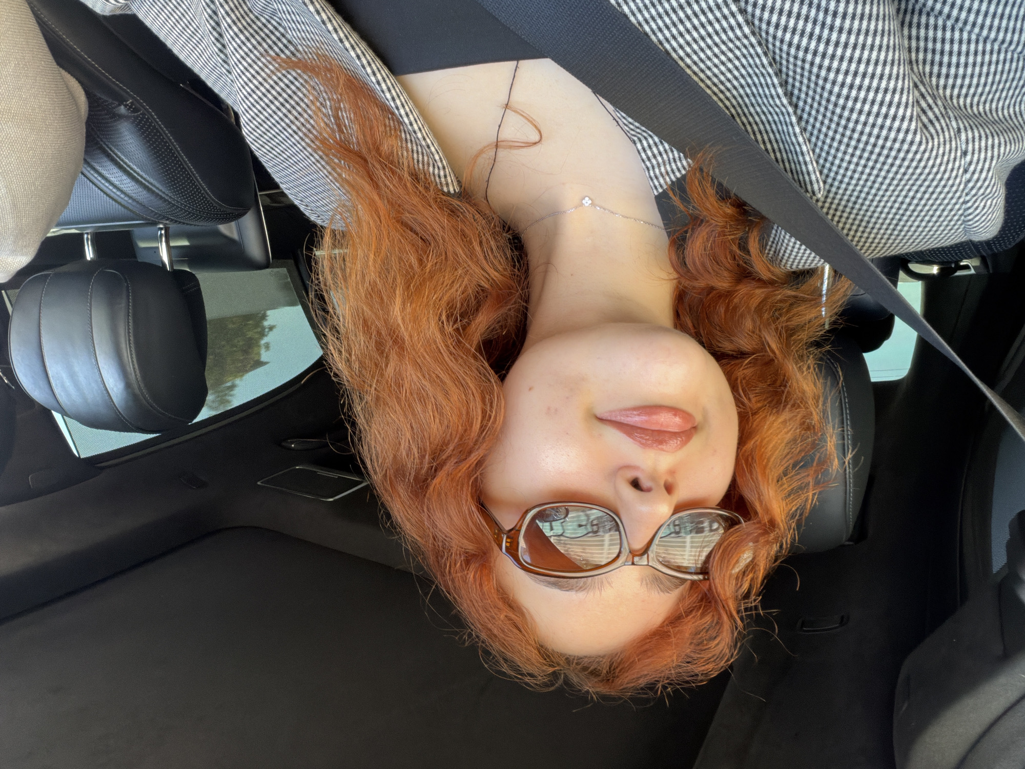 A person with auburn hair, wearing sunglasses, a black top, and a checkered blazer, is seated in a car with a seatbelt on. The car's interior includes black leather seats. The scene is not determinable as being in Strasbourg, France, from visual details alone.