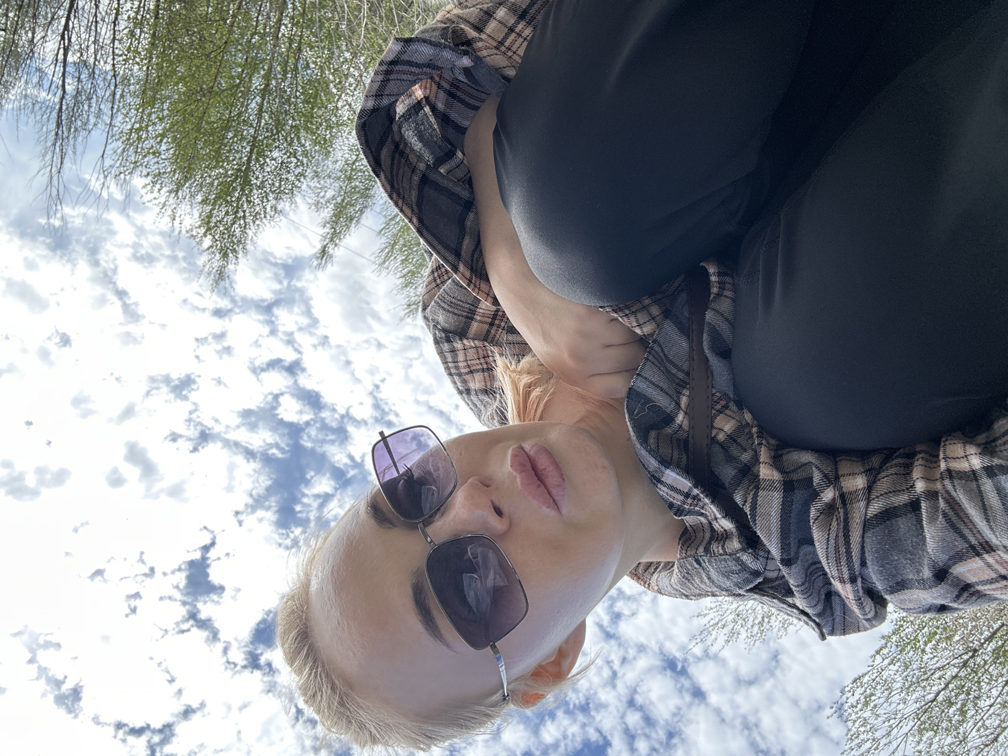 A low-angle, close-up shot shows a person with light skin, wearing sunglasses and a plaid shirt. They are dressed in black pants. The background includes a cloudy sky and tree branches with green leaves, suggesting an outdoor setting. The location is Высокий, Russia.