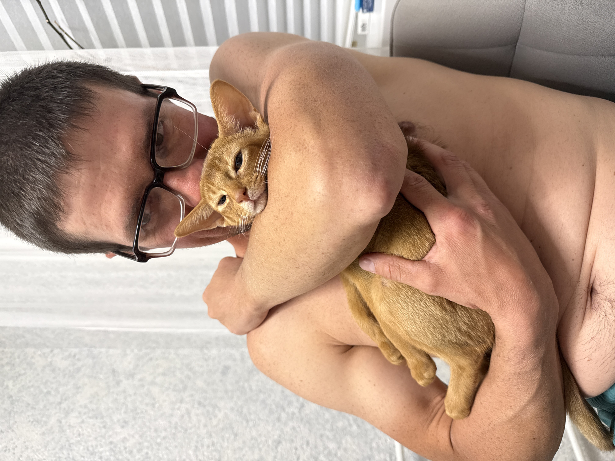 A shirtless man wearing glasses is holding a small, orange tabby cat. The man's arms are wrapped around the cat, and the cat is nestled against his chest. The man is looking down at the cat, and the cat is looking towards the camera with its eyes partially closed. The background is a plain wall and a window with blinds. The scene is indoors and appears to be in a home or similar private setting. The image does not contain any information about the city of Stary Oskol, Russia.