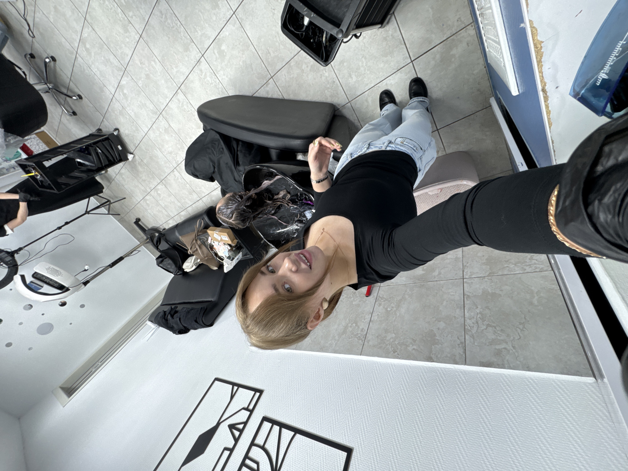 A young Caucasian woman with light brown hair, wearing a black long-sleeved shirt and light blue jeans, stands in a hair salon. She is taking a selfie, with her right arm extended towards the camera. She has on large beige earrings and a delicate necklace. Her makeup includes noticeable eyeshadow. In the background, a client is seated in a shampoo chair with their head tilted back, their hair wet and undergoing a treatment. A black hair dryer, a trolley with hair products, and other salon equipment are visible. The floor is tiled in a light grey. A black and white abstract artwork hangs on the wall to the left. The image was taken at a hair salon in the city of Chaikovsky, Russia.