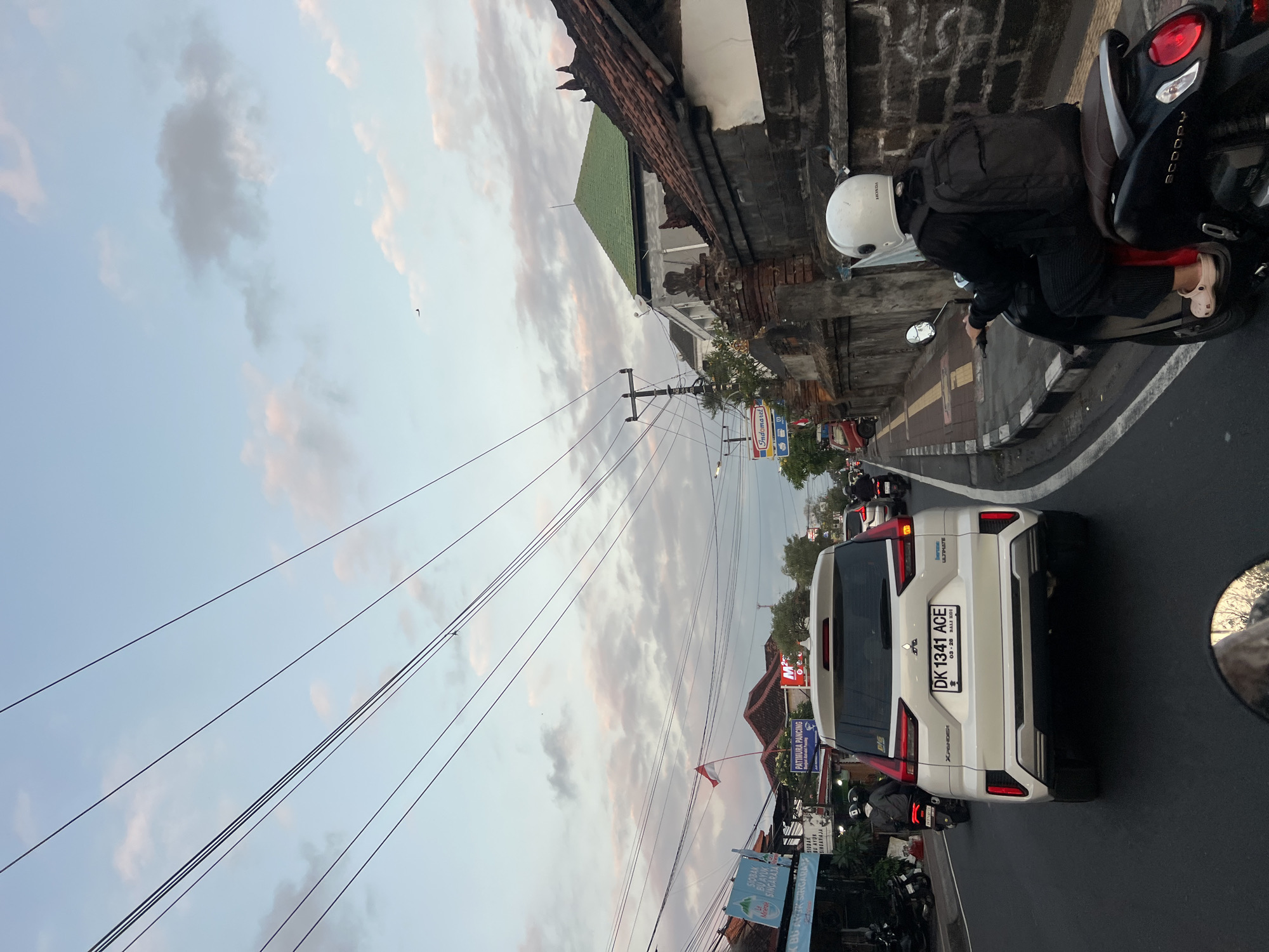 The image captures a street scene in Denpasar, Indonesia, likely during the late afternoon or early evening, given the soft, diffused light and pastel hues in the sky. The viewpoint is from behind a scooter, looking forward towards traffic on a paved road.
Dominating the center of the frame is the rear of a white Mitsubishi Xpander SUV, its license plate reading "DK 1341 ACE". To the right of the SUV, a person on a dark scooter, wearing a white helmet and a black jacket and pants, is visible. They are carrying a black backpack and have light-colored slip-on shoes.
The road is lined with buildings and structures typical of a Southeast Asian urban environment. On the left, storefronts with signage in Indonesian are visible, including "Indomaret". Further down the street, a flagpole with the Indonesian flag is flying. On the right, a traditional Balinese stone wall with intricate carvings runs alongside the sidewalk. Overhead, a dense network of electrical and telephone wires crisscrosses the sky.
The sky itself is a blend of pale blue and soft pink and gray clouds, suggesting either sunrise or sunset. A faint dark speck, possibly a distant bird or airplane, is visible in the upper portion of the sky. The overall atmosphere is one of a busy, everyday urban commute.