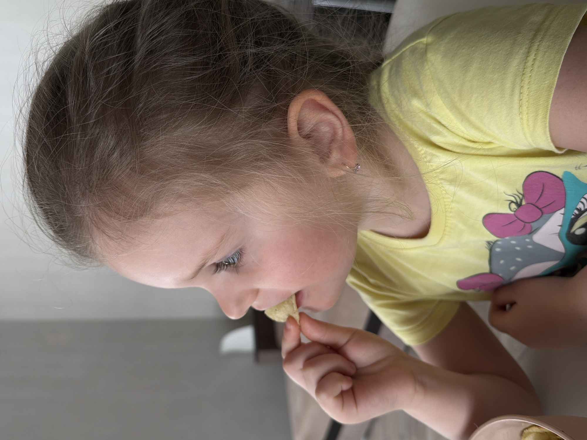 A young girl with light brown hair is eating a chip. She is wearing a yellow t-shirt with a cartoon character on it. The setting appears to be indoors, possibly in a home or dining area. It is not possible to determine the location to be Chaikovsky, Russia from the visual information provided.
