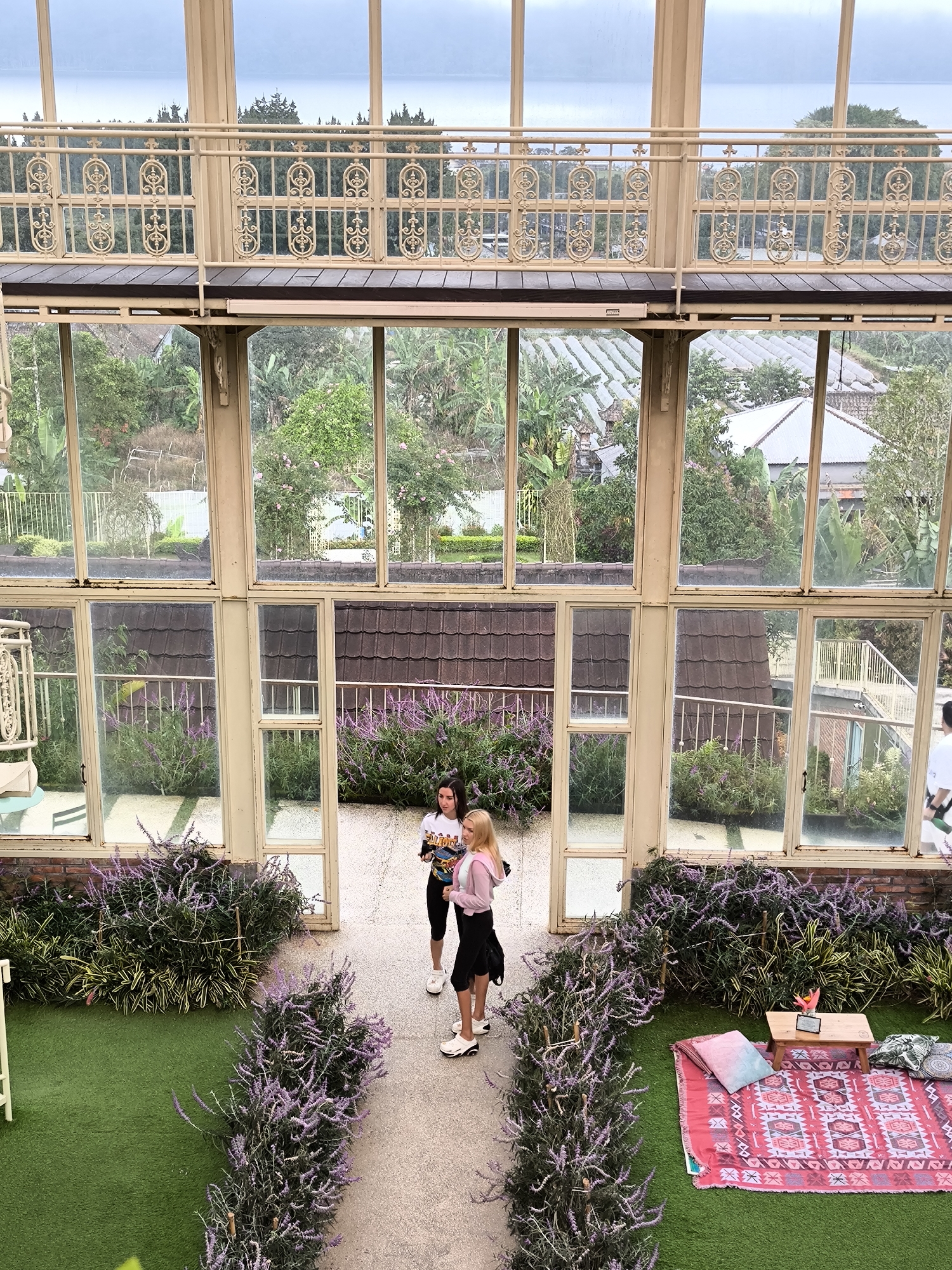 The image captures an elevated view of a garden area within a building or greenhouse-like structure, presumably located in Candikuning, Indonesia, given the presence of tropical foliage in the landscape visible through the windows.
Two young women stand in the center of a paved pathway lined with lavender bushes. One woman has dark hair and wears a white t-shirt, black shorts, and white sneakers; the other has blonde hair and wears a pink sweater, a black skirt, and white sneakers.
The path is flanked by artificial turf on either side, creating small garden sections. To the right of the path is a picnic setup on the turf. This includes a red patterned rug, several pillows, and a small wooden table with a vase of flowers.
The entire scene is framed by the structure of the building, which consists of large windows and ornate metal railings, giving a view of the surrounding lush landscape, including a tiered landscape with homes, trees, and cultivated fields. The upper portion of the structure has a balcony with ornate metal railings. The lighting appears to be overcast or diffused, which creates an even distribution of light throughout the scene.