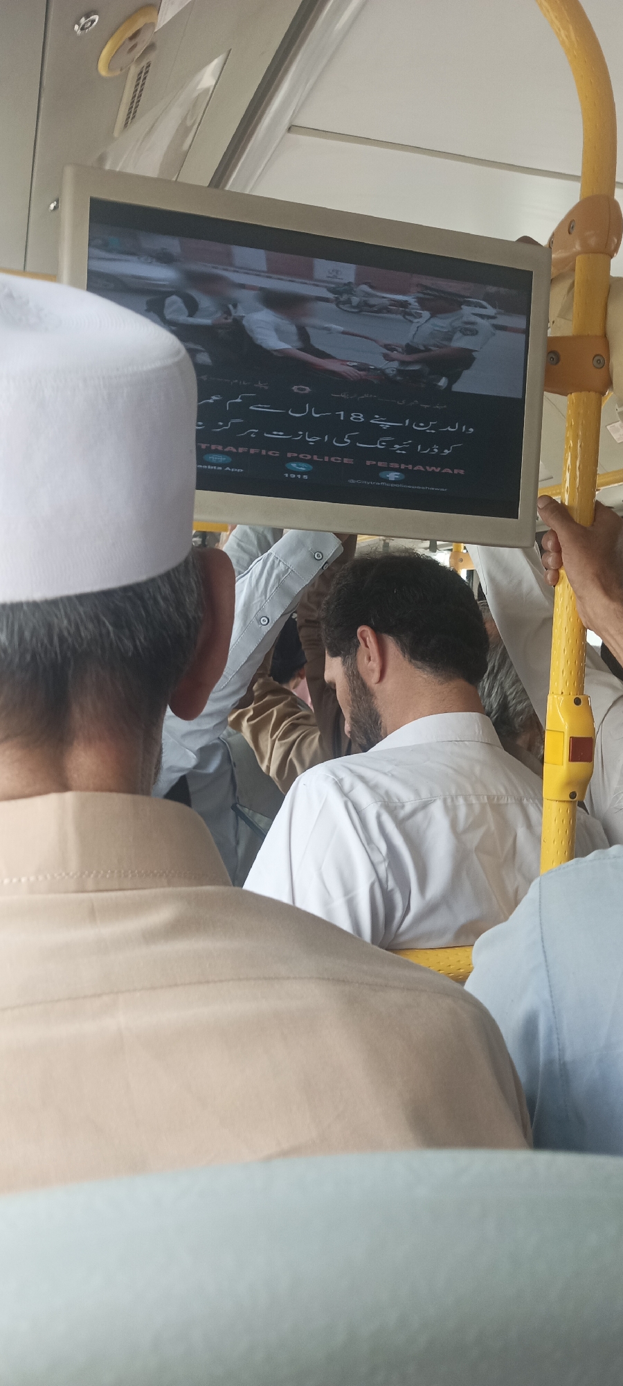 A screen displays a message in Urdu and English. The Urdu text reads: "والدین اپنے 18 سال سے کم عمر کو ڑا موٹر سائیکل کی اجازت ہرگز نہ دیں" which translates to "Parents do not allow their children under 18 to ride motorcycles". Below this, in English, it says "TRAFFIC POLICE PESHAWAR". Further down, there are logos for a "Saabta App" and "1915", along with a Facebook handle "@CityTrafficPolicePeshawar". The screen is mounted on the yellow pole of a vehicle, likely a bus, given the presence of passengers in the foreground. The people in the foreground, seen from behind, are wearing traditional clothing. The overall scene suggests a public awareness campaign about road safety for minors, disseminated in a public transport setting in Peshawar, Pakistan.