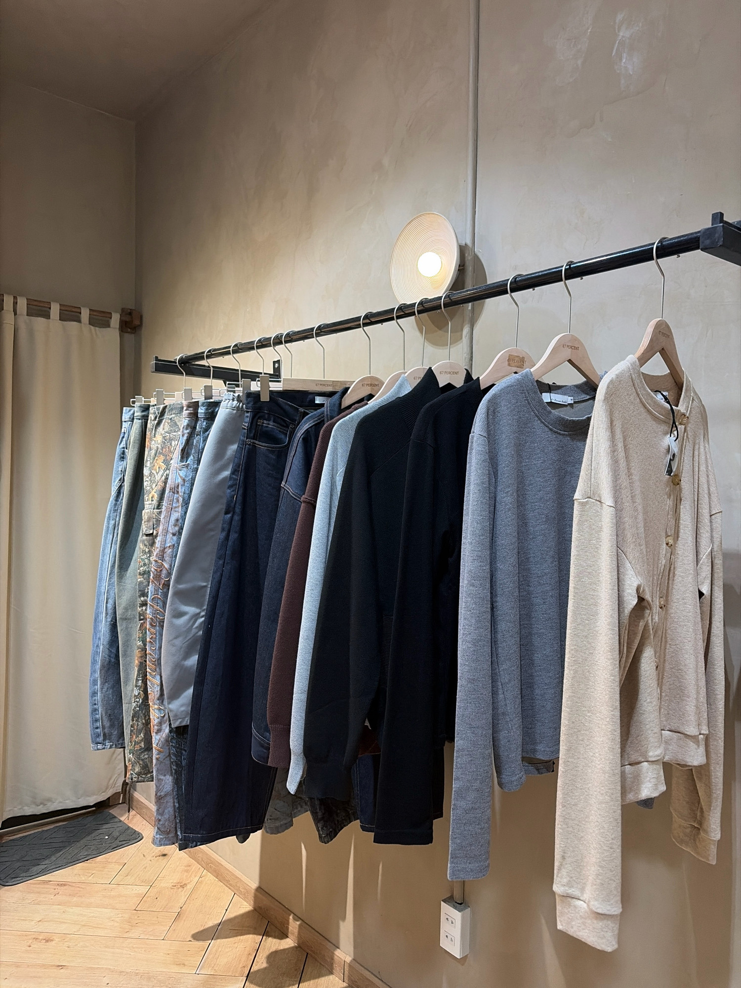 An indoor shot depicts a clothing rack filled with various garments. From left to right, the items include: denim pants, patterned pants, light gray pants, dark blue jeans, a dark brown jacket, a gray long-sleeved shirt, and a beige cardigan. The clothing rack is supported by black metal brackets against a textured, light brown wall. A round, white light fixture is mounted on the wall above and to the left of the clothing rack. To the left of the clothing rack, a cream-colored curtain hangs, partially obscuring a doorway. The floor is made of light brown wooden planks laid in a herringbone pattern. A dark gray doormat is positioned on the floor in front of the curtain. A white electrical outlet is visible on the wall below the clothing rack. The scene appears to be set in a retail environment or a personal wardrobe. The image is taken at eye-level, with the camera focused on the clothing and the wall. There are no people visible in the image. This scene is not in Da Nang, Vietnam.