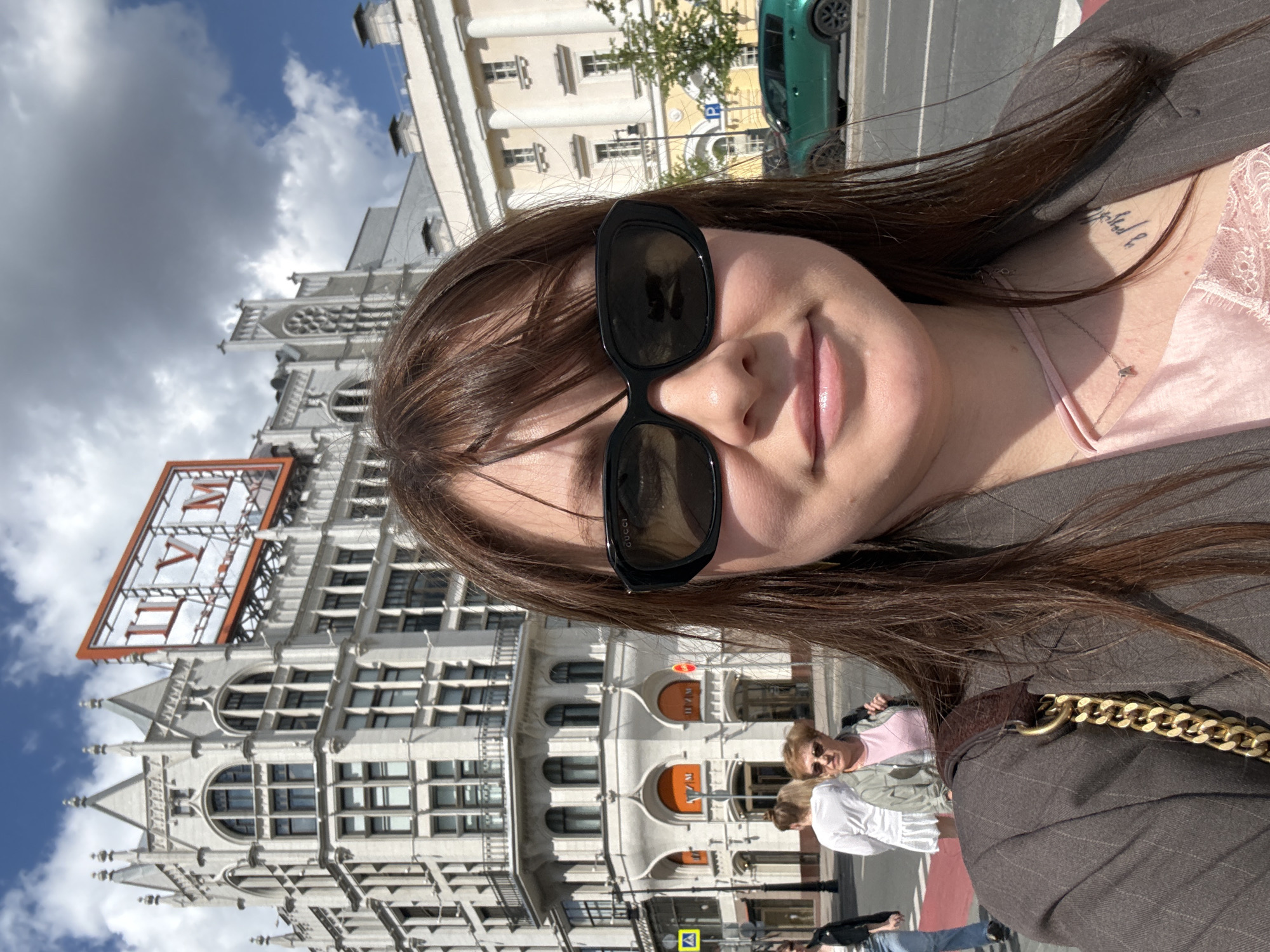 A selfie-style portrait captures a young woman outdoors in a city setting, likely Moscow, Russia, as indicated by architectural cues and specific signage. The image is taken from a slightly low angle, showing the person's upper body and a bustling street scene in the background.
The main subject, a woman with dark brown hair, is wearing black sunglasses, a light pink top, and a dark pinstripe blazer. She has a subtle smile on her face. A delicate necklace and a tattoo with script text on her right collarbone are visible. The word "GUCCI" is faintly discernible on the arm of her sunglasses.
The background features several ornate, multi-story buildings with intricate architectural details, including arched windows and decorative elements. A prominent feature is a large, square sign or clock face on a building, displaying the Roman numerals "VII" and "IX" separated by the Cyrillic letter "И" (meaning "and"). This sign strongly suggests the Central Telegraph building in Moscow. Another building facade exhibits distinctive orange arched entrances.
On the street, blurry figures of other pedestrians, mostly women, are visible, along with a green car parked further down the street. A blue "P" parking sign and a yellow square sign with a black arrow are also present. The scene is illuminated by bright daylight, with a clear blue sky and some white clouds, indicating sunny weather. The overall impression is a vibrant urban environment.