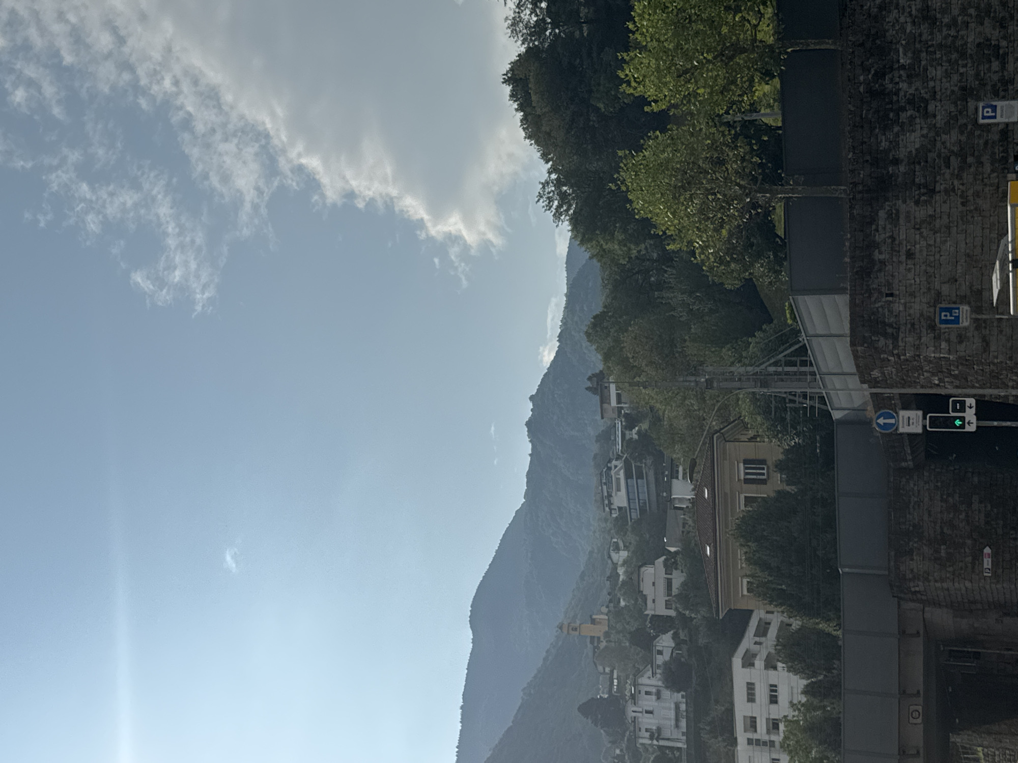 The image shows a view of houses nestled on a hillside in Bellinzona, Switzerland, with a mountain range in the background. A bright, hazy sun illuminates the sky, which has scattered clouds. In the foreground, there is a dark, brick-lined tunnel opening. Above the tunnel, a grey wall and fence are visible, with trees and buildings behind them. A traffic light with a green signal is also present. The architecture of the houses suggests a European style, with a prominent yellow building with a steeple visible further up the hill, likely a church. The overall scene depicts a residential area integrated into a natural, mountainous landscape.