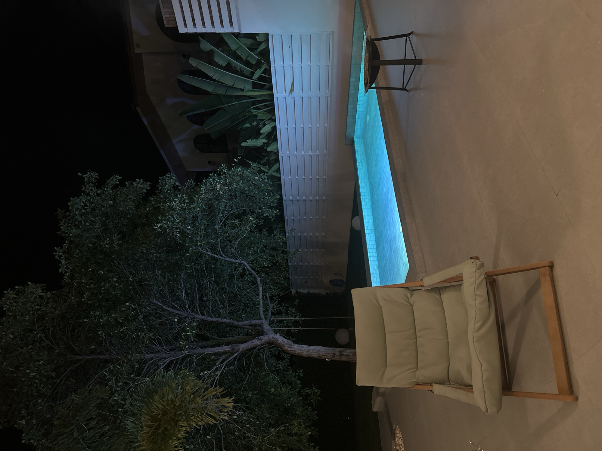 This is a medium shot, full-frame photograph taken at night in the vicinity of ตำบลเกาะพะงัน, Thailand, depicting a tranquil outdoor recreational area. In the foreground, a light green, padded lounge chair made of wood and fabric sits on a light-colored tiled patio. To the right of the chair, a small, dark metal side table with a tripod base holds what appears to be a fire pit. Beyond the patio, a bright blue, illuminated swimming pool occupies the middle ground, its water emitting a vibrant glow. The pool is bordered by a low concrete wall. Behind the pool, a white horizontal slatted fence runs across the width of the image, separating the pool area from the background. A mature tree with a thick trunk and spreading branches, draped with what looks like a swing, stands to the left of the pool. Lush green foliage and a palm frond are visible behind the tree. In the far background, a beige building with arched windows and a sloped roof is partially obscured by large, dark green banana leaves. The scene is illuminated by artificial lighting, creating a serene and inviting atmosphere.