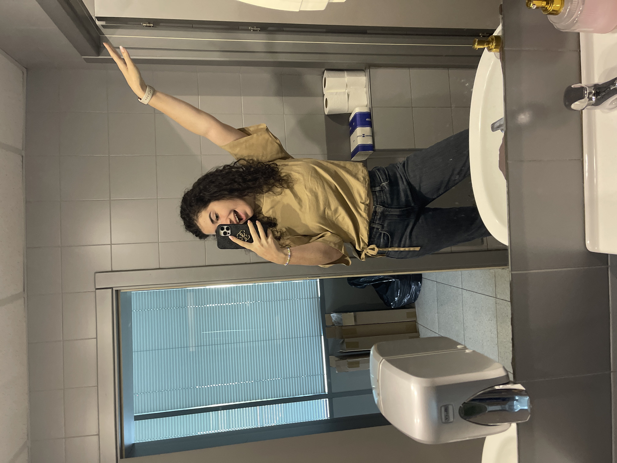 A young woman with curly hair is taking a selfie in a bathroom mirror. She is wearing a tan top and dark jeans. Her arm is raised.
The bathroom has gray tiled walls, a white sink, a hand dryer, and a window with blinds. Toilet paper and a dispenser are visible on the wall. Several cardboard boxes are visible behind the window.
I am unable to determine the exact location within Sofia, Bulgaria.