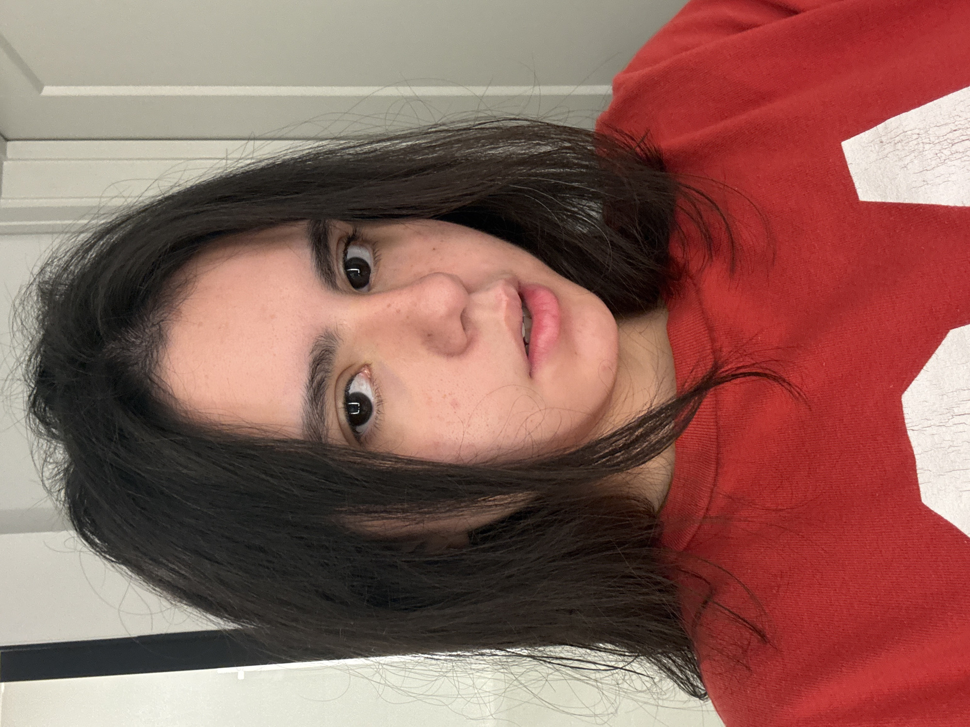 A woman with dark brown hair and brown eyes is wearing a red sweatshirt with a white design. She is positioned in front of a white door frame. There is no indication of location in Austin, Texas.