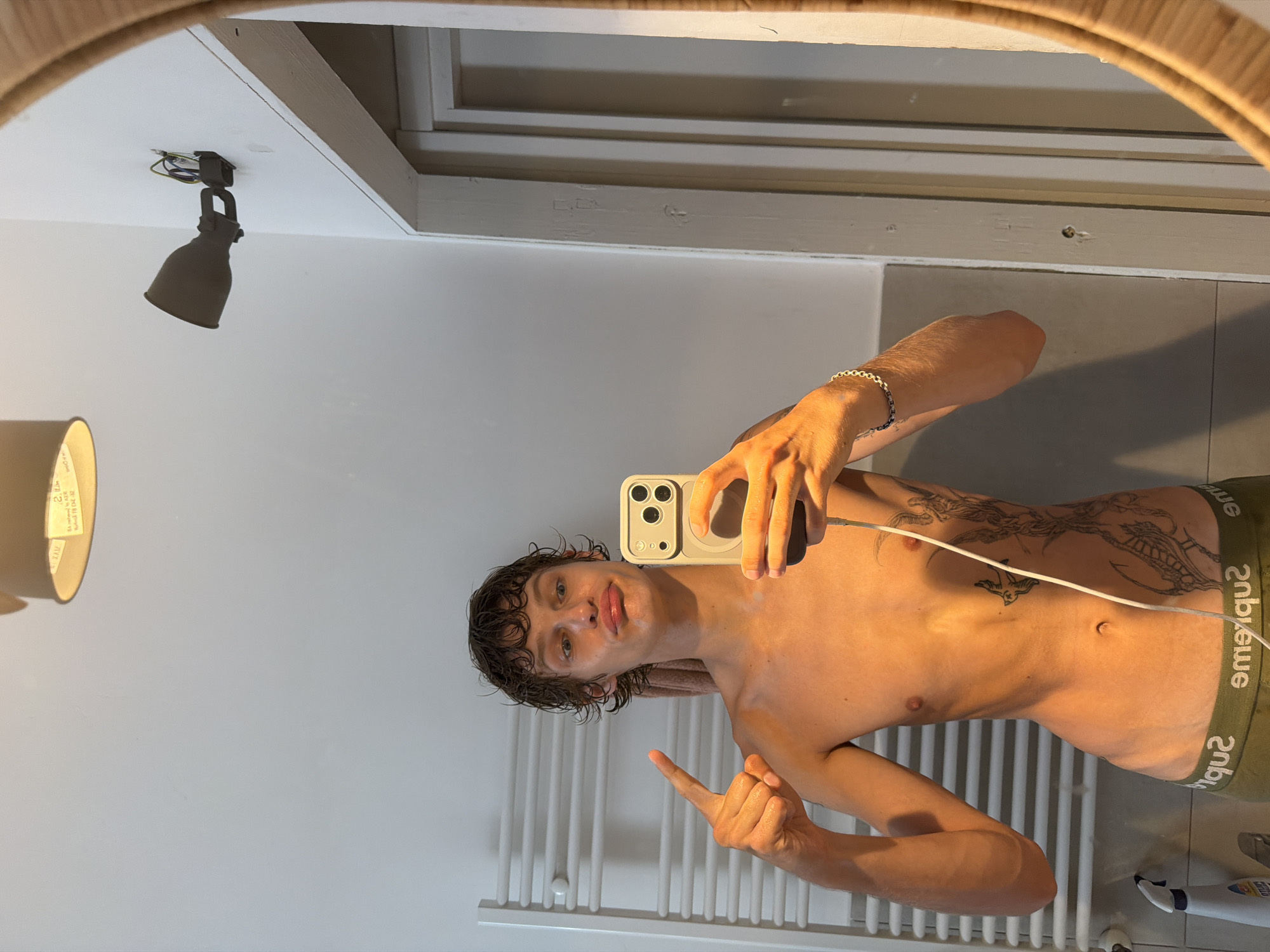 A young man with curly brown hair and a slender build, shirtless and wearing olive green boxer briefs, stands in a bathroom and takes a mirror selfie with his phone. He is making a puckered face and pointing his left index finger towards the camera. His torso is adorned with a large black ink tattoo on his right side, depicting what appears to be a dragon or serpent. To his left, a white heated towel rack is visible. The bathroom features gray tiled walls and a light fixture on the ceiling. The overall lighting suggests it is daytime. There is no indication of the specific location in Milan, Italy.