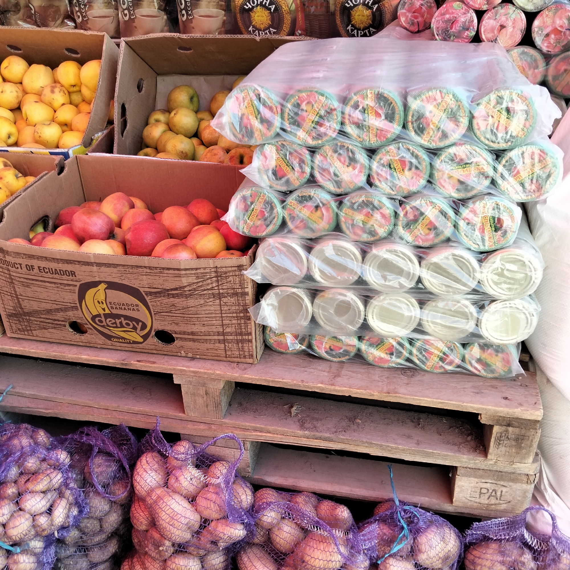 The image depicts a street market scene in Cupcini, Moldova, with a focus on various food items displayed for sale. The foreground features sacks of potatoes, some in purple netting and others in lighter colored netting. Behind the potatoes, stacked wooden pallets support several cardboard boxes. One box, labeled "PRODUCT OF ECUADOR," contains red and yellow apples. Another box, also containing apples, is visible to the left.
Further back, a stack of individually wrapped items, possibly cups or lids, is covered in plastic. The wrappers have colorful designs and some Cyrillic text, suggesting they are intended for the local market. In the background, bags of coffee or similar products are visible, also with Cyrillic lettering.
The scene suggests a daytime market, likely during business hours. The overall impression is one of commerce and fresh produce available to consumers. There are no people visible in the image, focusing entirely on the goods for sale and their arrangement. The presence of produce from Ecuador alongside local items indicates trade and a diverse offering at the market. The visible text includes brand names like "derby" and "EPAL" on the packaging and pallets, as well as product information in Russian.