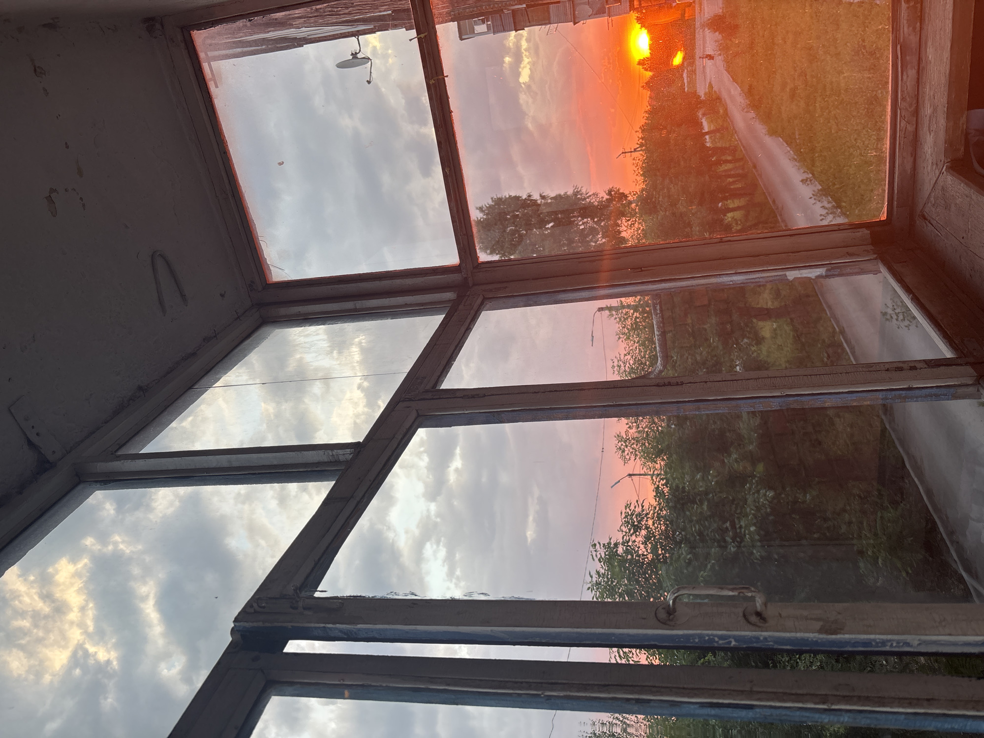 The image captures a sunset viewed through a multi-paned window frame. The window panes are dirty and the wooden frames are weathered, suggesting an older building. The sky is dominated by a vibrant sunset, with hues of orange, pink, and yellow. Clouds are present, some illuminated by the setting sun.
In the top right pane, a bright sun is visible on the horizon, casting a warm glow over the scene. A road or path runs through a grassy area alongside trees. The reflection in the glass shows trees and possibly buildings, indicating an outdoor setting. A satellite dish is visible on the exterior wall of the building in the top left pane.
The lower panes reflect more of the sky and greenery, with a hint of a streetlamp or utility pole visible in the distance in one of the reflections. The overall impression is one of a peaceful, late evening scene in a semi-urban or rural environment. The golden hour lighting creates a serene and slightly melancholic mood. No people are visible in the image. There is no visible text.