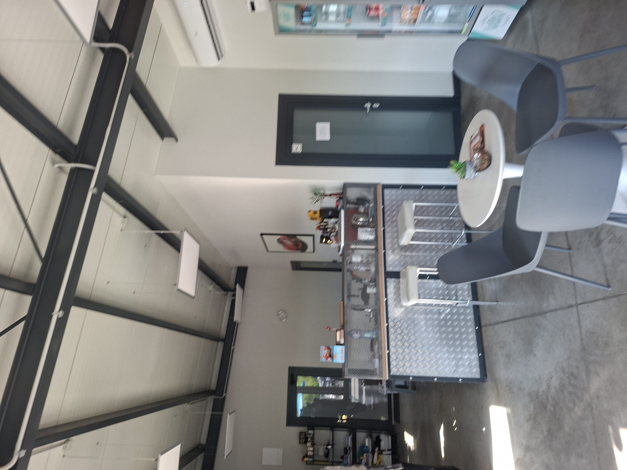 The image depicts an interior space with a modern, industrial aesthetic. The walls are painted in a light grey tone, contrasted by a black structural framework, possibly indicating a warehouse or showroom environment in Unknown, Unknown. Natural light enters through a front window, as well as other windows and doors in the back, where shelves of products can be observed.
The floor appears to be a polished concrete surface. A small, round table is positioned near the front, accompanied by three grey chairs, indicating a casual meeting or waiting area. A plant sits on the table's surface.
Along the wall is a counter or display unit with a metallic, industrial-style design. The counter area features a tiered structure and small stools. Decorations can be seen along the countertop.
Overhead, fluorescent lights are suspended from the structural framework. Also overhead are an air conditioning unit and a shelving unit. A picture hanging on the wall in the background is visible. The overall environment suggests a blend of retail and industrial functionality.
