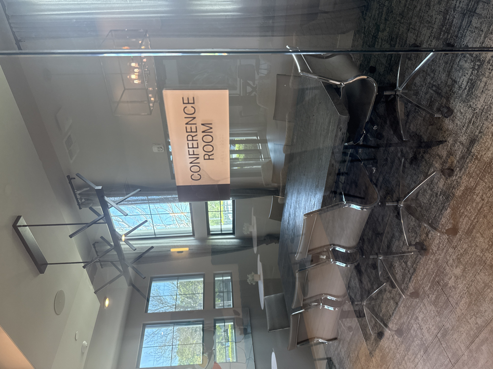 This forensic image depicts the interior of an unoccupied modern conference room, located in Orlando, United States, viewed through a reflective clear glass partition. A prominent rectangular sign on the glass states "CONFERENCE ROOM" in black text, with supplementary smaller text and braille underneath. The room contains a large, dark-finished rectangular conference table. Eight contemporary office chairs with light-colored seating surfaces and chrome five-star bases on casters are arranged around the table; six are clearly visible. Overhead, two distinct chandeliers illuminate the space: a linear, abstract metal fixture positioned directly above the conference table, and a larger, rectangular cage-style chandelier located further into the room. Recessed ceiling lights and a circular ceiling speaker are also present. The left wall is dominated by multiple large windows, offering a partial exterior view of green trees under natural daylight. Light-colored curtains are drawn along the right side of the room. The flooring is a dark, patterned carpet. Beyond the main conference area to the left, two small, round white pedestal tables, each adorned with a white floral arrangement, are visible. No individuals are present, and no actions are occurring within the frame.