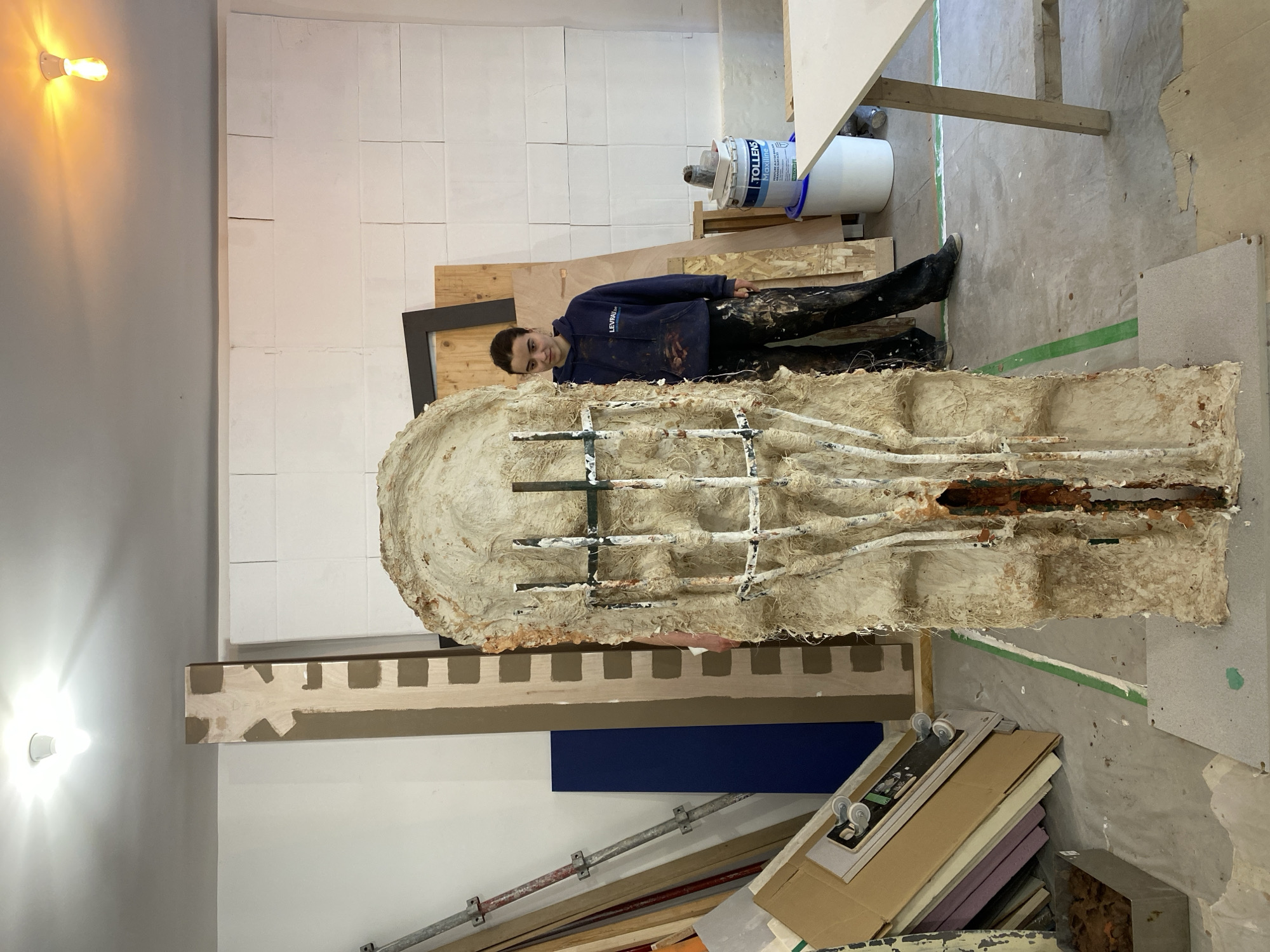 A horizontal, high-angle indoor photograph shows a young man standing to the right of a large, vertical, irregular-shaped sculpture, possibly in a studio or workshop. The man, facing the camera with a slight smile, is dressed in a dark blue hooded sweatshirt with the word "LEVRILL" printed on it, dark pants, and dark shoes. His clothing is splattered with what appears to be paint. He is positioned slightly behind the sculpture, appearing to lean against it.
The sculpture itself is a complex structure. It has an outer layer of beige-colored material, possibly plaster or clay, and an internal framework of metal rods and pipes. There is a dark orange-brown section in the center of the sculpture.
The background consists of a wall constructed of white blocks, possibly concrete blocks, and various pieces of plywood and lumber. To the right of the man, there is a large white bucket and a table with a light-colored wooden top.
The foreground contains various materials, including stacked cardboard boxes, foam, a piece of dark blue paneling, and metal pipes or supports. A bare light bulb is visible in the upper left corner of the image.
There is no indication in the image that the location is in Paris, France. The scene appears to be a workshop or studio, possibly an artist's workspace.