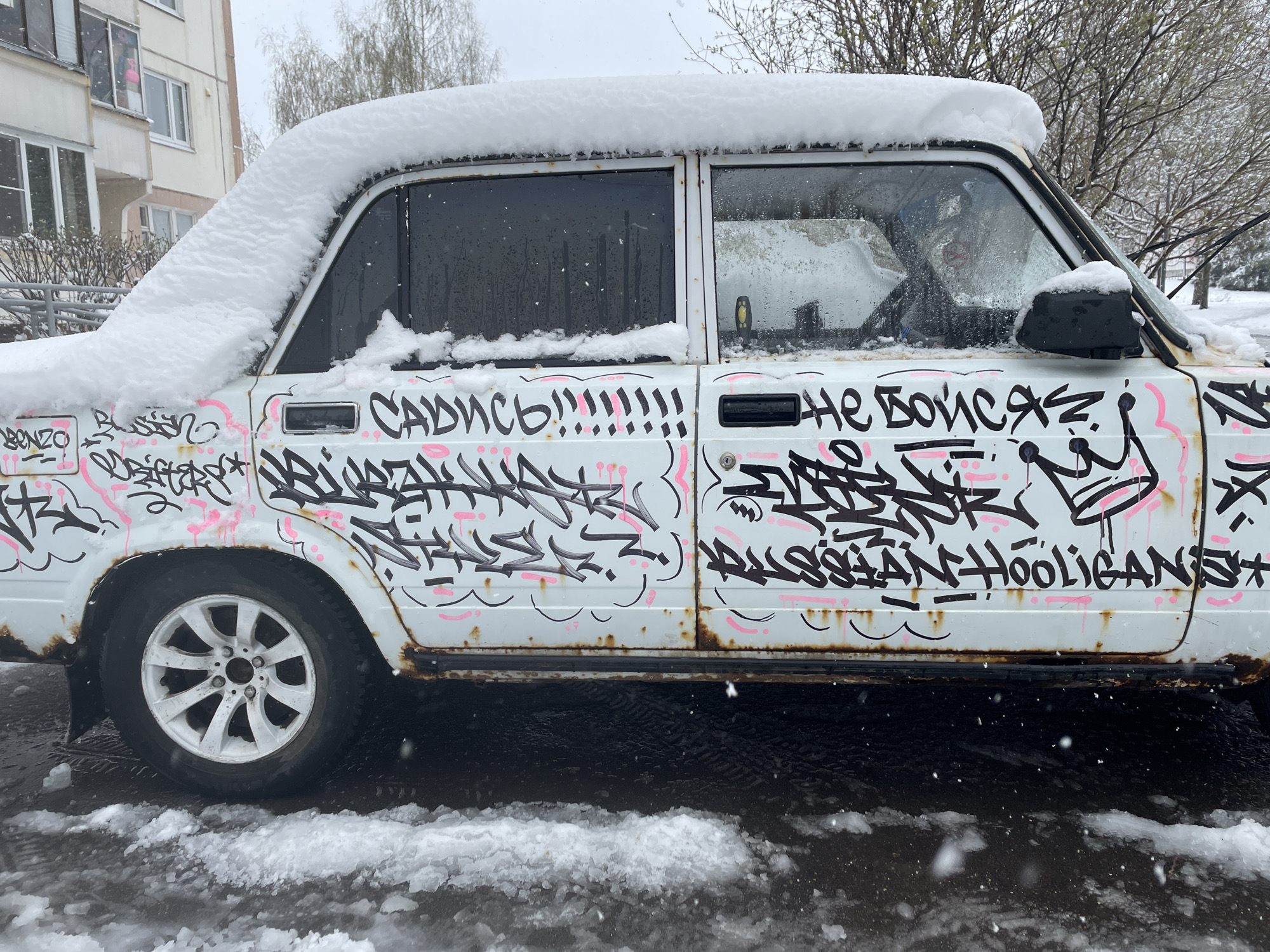 An old, white sedan, likely a Lada, is parked outdoors during a snowy day in what appears to be Moscow, Russia. The car is heavily covered with graffiti in black and pink spray paint, contrasting with its rusted lower body and door seams. A thick layer of snow blankets the roof, hood, and window frames, while light snowflakes are visible in the air and on the car's windows.
The graffiti features stylized lettering and abstract designs. Visible text includes:
- On the front door: "САДИСЬ!!!!!!!!!" (SADIS'! - meaning "Get in!" or "Sit down!"), and "BONZO".
- On the back door: "НЕ БОЙСЯ" (NE BOYSYA - meaning "Don't be afraid") and "RUSSIAN HOOLIGANS" with a crown symbol.
The car's wheels are white multi-spoke rims. The ground beneath the car is a mix of wet asphalt and melting snow. In the background, to the left, is a beige apartment building with multiple windows, some open. Bare trees and branches are visible behind the car, suggesting winter or early spring. The sky is overcast, and the overall atmosphere is cold and urban. No people are visible.