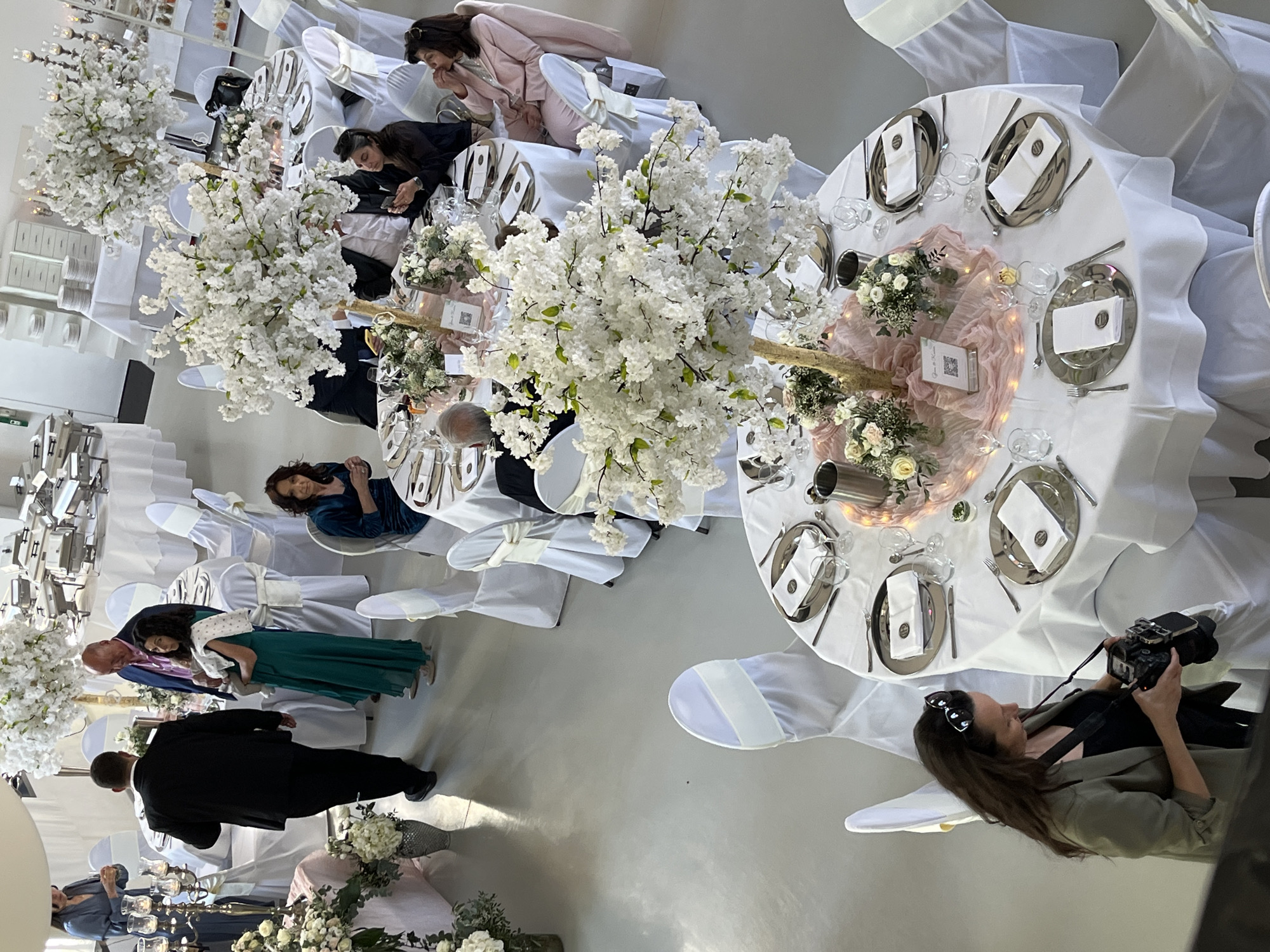 The image captures an indoor event, likely a wedding reception or a formal gathering, in Saarbrücken, Germany. The scene is viewed from a slightly elevated perspective, looking down onto several round tables set for dining.
The room features white tablecloths and chairs adorned with white covers and sashes, creating a bright and elegant atmosphere. Each table is meticulously set with silver charger plates, white folded napkins bearing a small black circular emblem, clear glassware, and silverware. Dominant centerpieces consist of tall, artificial white cherry blossom trees on golden stands, complemented by smaller floral arrangements of white and light pink flowers with greenery. Pink ruffled fabric runners, subtly illuminated by fairy lights, and small votive candles enhance the festive decor. A small card with a QR code and partially visible text, possibly "Gäste haben," is placed on one table.
Several guests are visible throughout the spacious venue. Some individuals are seated at the tables, dressed in formal or semi-formal attire, engaged in conversation or looking at their phones. Notable figures include a woman in a pink pantsuit and another in a blue velvet dress seated at tables, a man in a blue suit and a woman in a dark green dress standing near a table, and a man in a black suit with his back to the camera. In the foreground on the right, a woman with dark hair and sunglasses perched on her head is holding a DSLR camera, appearing to capture moments of the event. The bright indoor lighting suggests a daytime or early evening event. In the background, white sideboards with stacks of plates and chafing dishes indicate a food service area. The overall mood appears calm and celebratory.