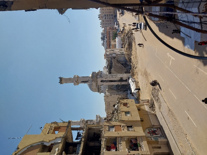 The image captures an urban street scene in Khan Yunis, Palestinian Territory, under a clear, bright blue sky. A light-colored mosque with a tall, slender minaret and a dome, appearing structurally damaged, dominates the midground. To the left, a multi-story building exhibits extensive damage, with large sections of its facade missing, exposing internal floors and structural elements. Laundry is visible hanging on balconies of a less damaged portion of this building, which also features graffiti or a mural on its lower wall. The paved street in the foreground shows faded white lane markings and a single turning arrow, with scattered debris. Piles of rubble and partially collapsed structures are visible throughout the scene, particularly around the mosque and other buildings. Several pedestrians walk on the right side of the street and sidewalks. A person operates a motorcycle in the midground, moving away from the viewer, and a white sedan is present further down the street on the right. Temporary shelters or stalls are discernible on the far right foreground.