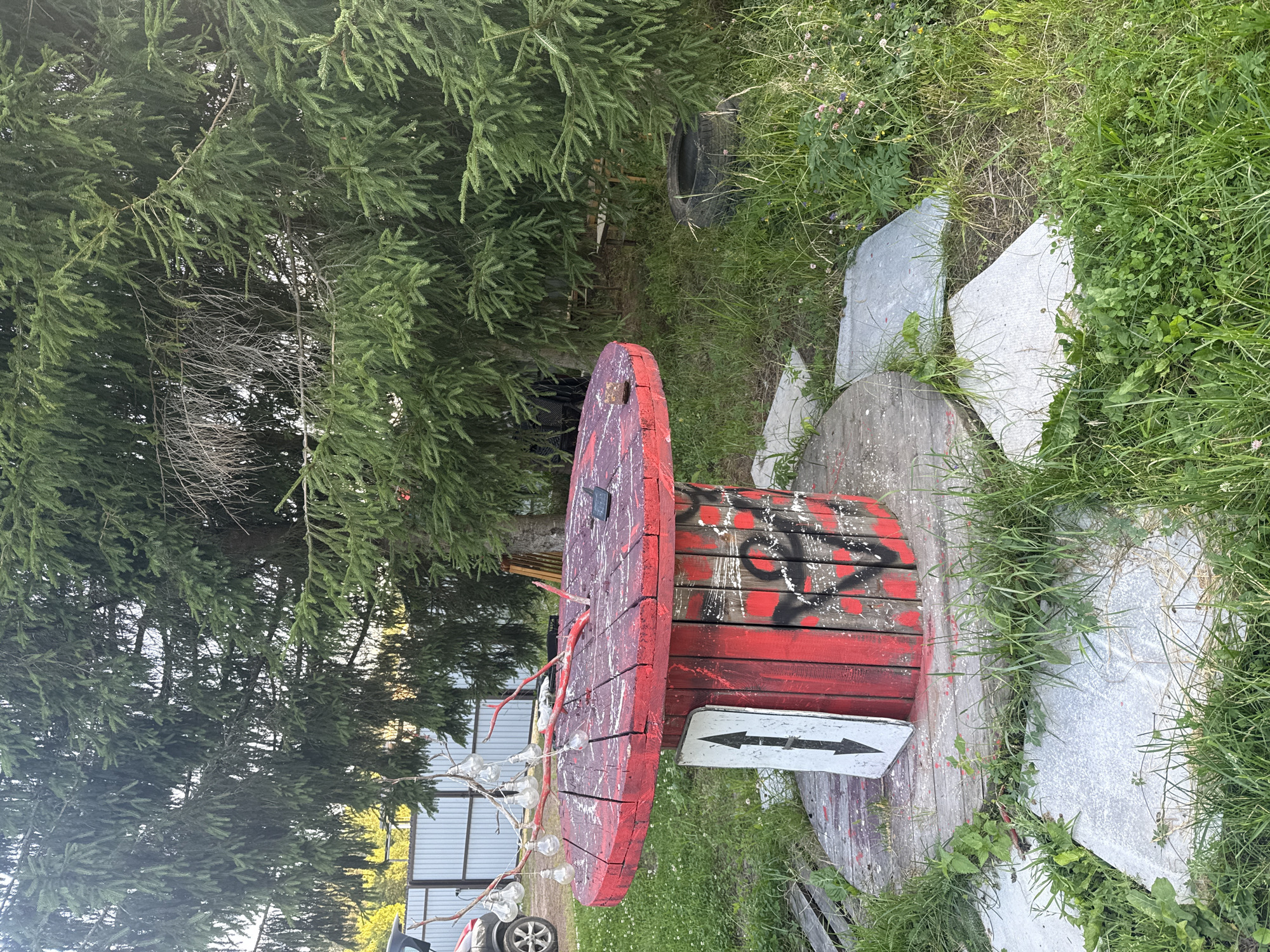 In the city of Lyady, Russia, an outdoor scene features a repurposed red cable spool table with graffiti. The table is adorned with a string of bare light bulbs hanging from branches, with some bulbs illuminated. A black smartphone rests on the table's surface. A white sign with black arrows pointing in opposite directions is placed beside the table. The area around the table is covered with grass and interspersed with large, irregular concrete slabs. In the background, a large evergreen tree dominates the landscape, with a section of a metal fence and a vehicle visible to the left. To the right, a discarded tire lies in the overgrown grass.