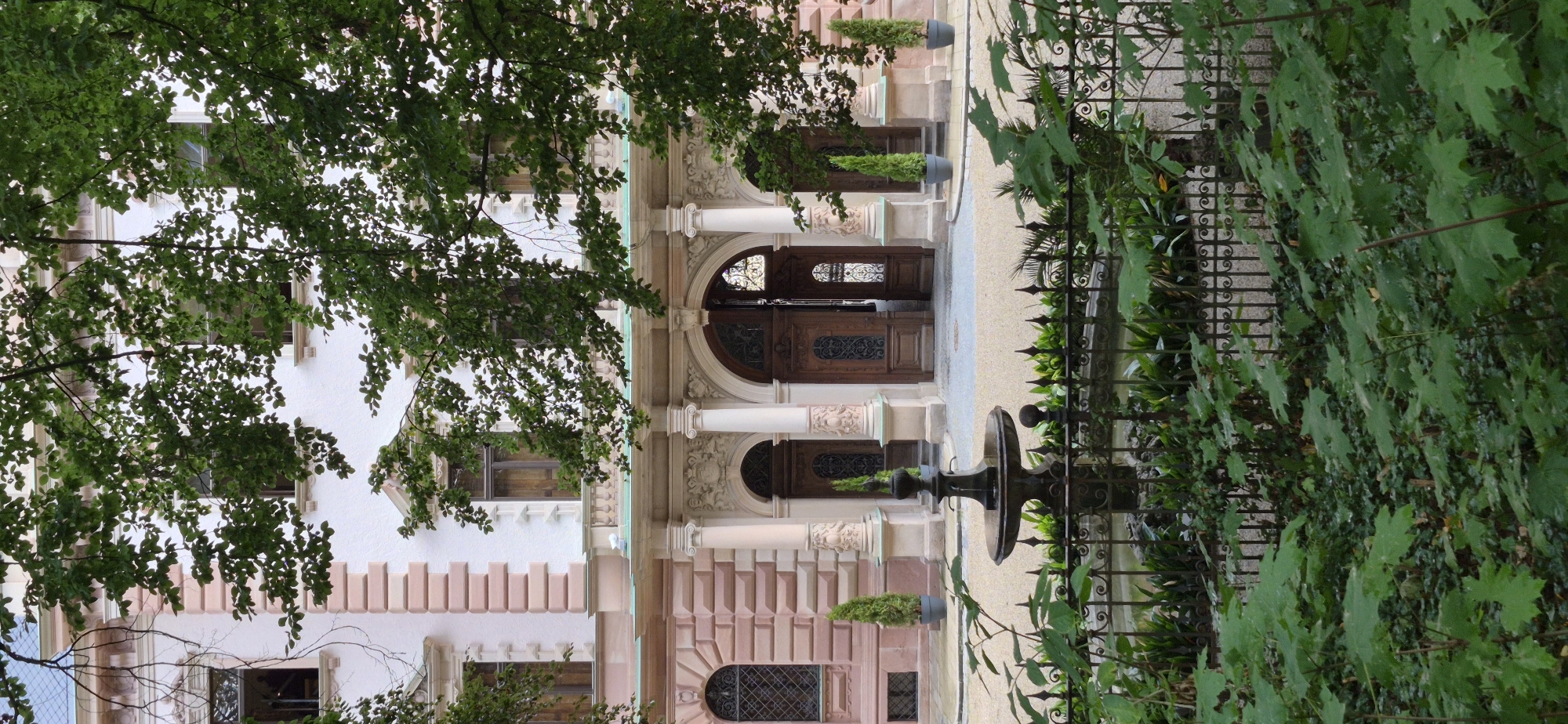 The image shows the facade of a pink and white building in Regensburg, Germany, with trees in the foreground. The building appears to be an old structure with classical architectural elements, including arched doorways, columns, and ornate carvings. A small fountain is visible in the courtyard in front of the building, with a decorative wrought-iron fence surrounding it. The courtyard is paved with gravel. The scene is dominated by the lush green foliage of trees, which partially obscure the view of the building and create a sense of depth and privacy. The overall impression is one of a serene and historic setting.