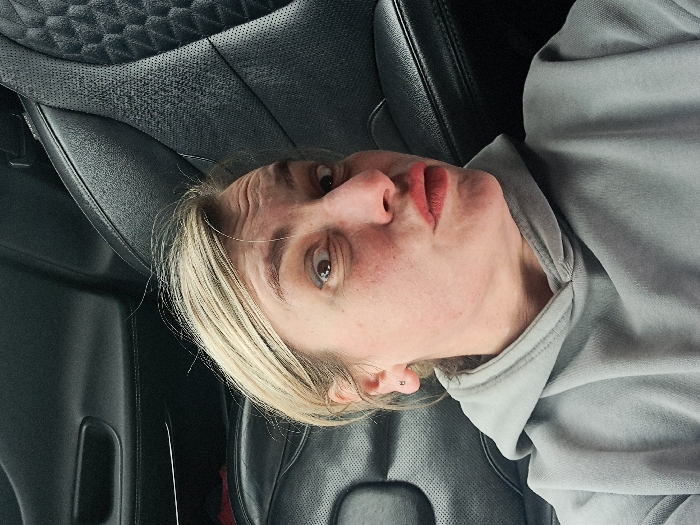 An adult female is depicted from the chest up, positioned within the passenger compartment of a vehicle. She has blonde hair, a light complexion with visible freckles, and wide, dark-rimmed eyes. Her eyebrows are raised, causing horizontal creases on her forehead, and her lips are pursed and tinted reddish, forming a "duck face" expression. A small, silver-colored stud earring is present in her left earlobe. She wears a light grey hooded sweatshirt with a visible drawstring at the neckline. The background consists of dark-colored, likely black, leather car upholstery, featuring a quilted or diamond-stitch pattern and perforated sections, consistent with a vehicle interior. A dark car door panel with a recessed handle is visible to the left. The specific geographic location within the city of Sasovo, Russia, cannot be determined from the visual information presented in the image.