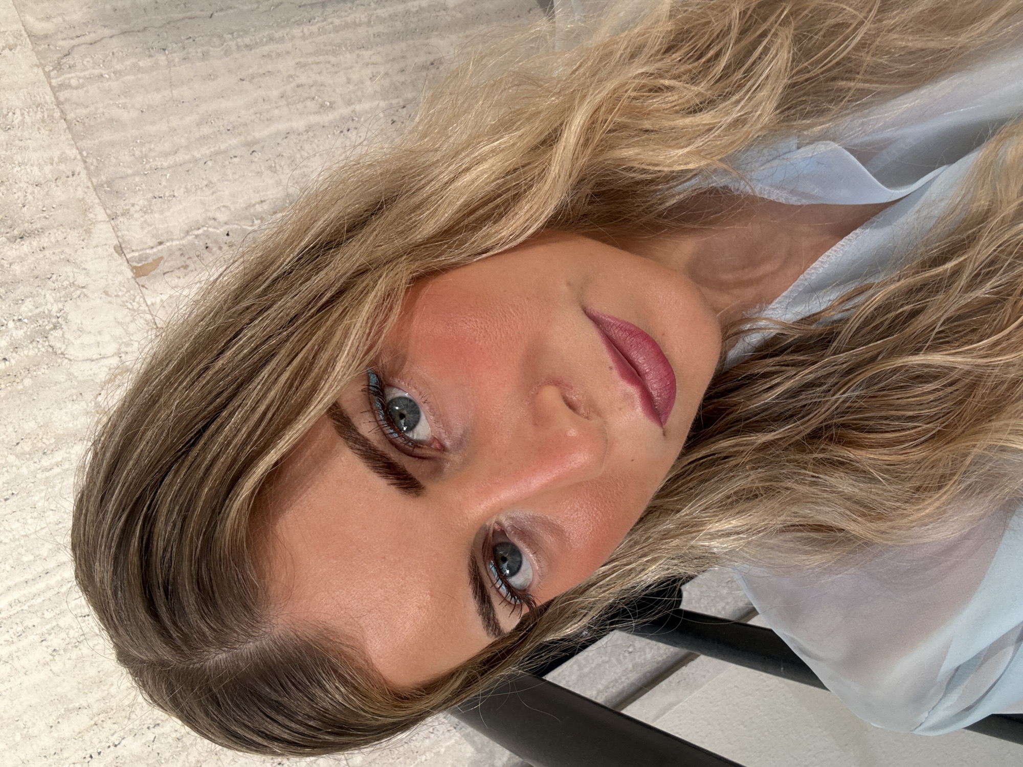 A close-up, rotated image captures a woman with blonde, wavy hair and fair skin. She has prominent cheekbones, full lips with dark lipstick, and light blue eyes. She appears to be wearing a light blue garment. The background includes a neutral-toned, textured surface, possibly tile or stone flooring, and a glimpse of a black object that could be a piece of furniture or a frame. The image composition emphasizes her facial features. The location in Paris is not determinable from the image.