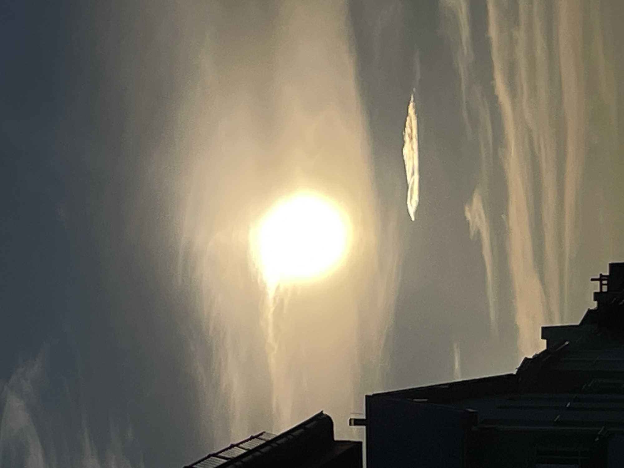 A bright, diffused sun is partially obscured by thin clouds, casting a warm, golden light on the sky. The sun is the focal point of the image, creating a luminous halo. Below the sun, a single, elongated cloud with a distinctly flat, layered appearance floats in the atmosphere. The sky transitions from a deep blue at the top to a lighter, warmer hue towards the horizon. Silhouetted buildings with visible architectural details, including a railing on a balcony or roofline, are present on the left side of the frame. The buildings are dark and their features are indistinct, suggesting they are viewed against the bright sky. The overall scene depicts an urban landscape under a bright, cloudy sky, likely during sunrise or sunset.