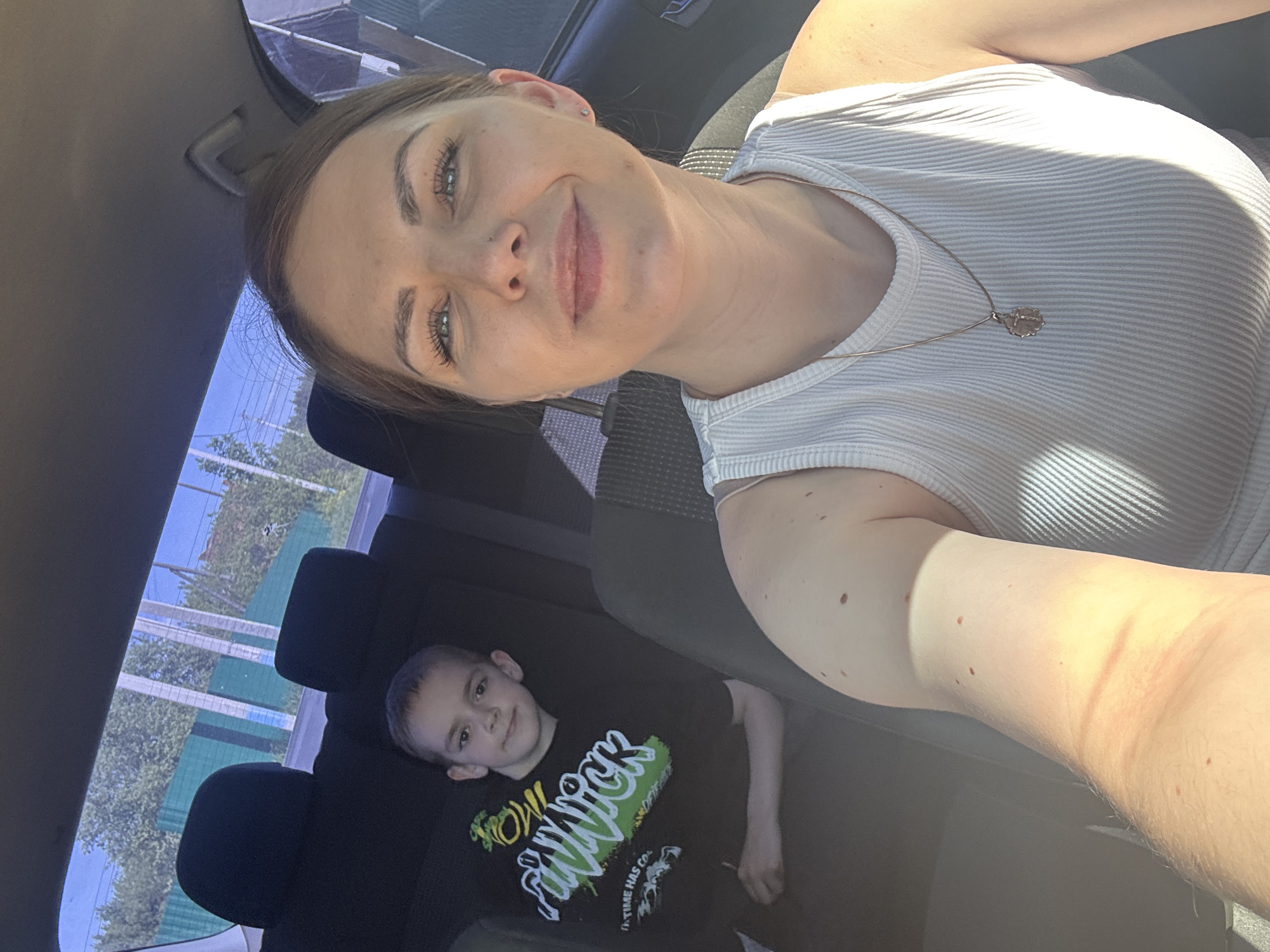 A medium shot depicts a woman and a child inside a car. The woman, likely in the driver's seat, is fair-skinned and wearing a white ribbed tank top with a gold necklace. Her arm is visible in the foreground, angled towards the camera. The child, in the back seat, is wearing a black t-shirt with a green graphic design. The background, visible through the car window, appears to show trees and a fence or barrier. The location is Razdolie, Russia, based on the prompt.