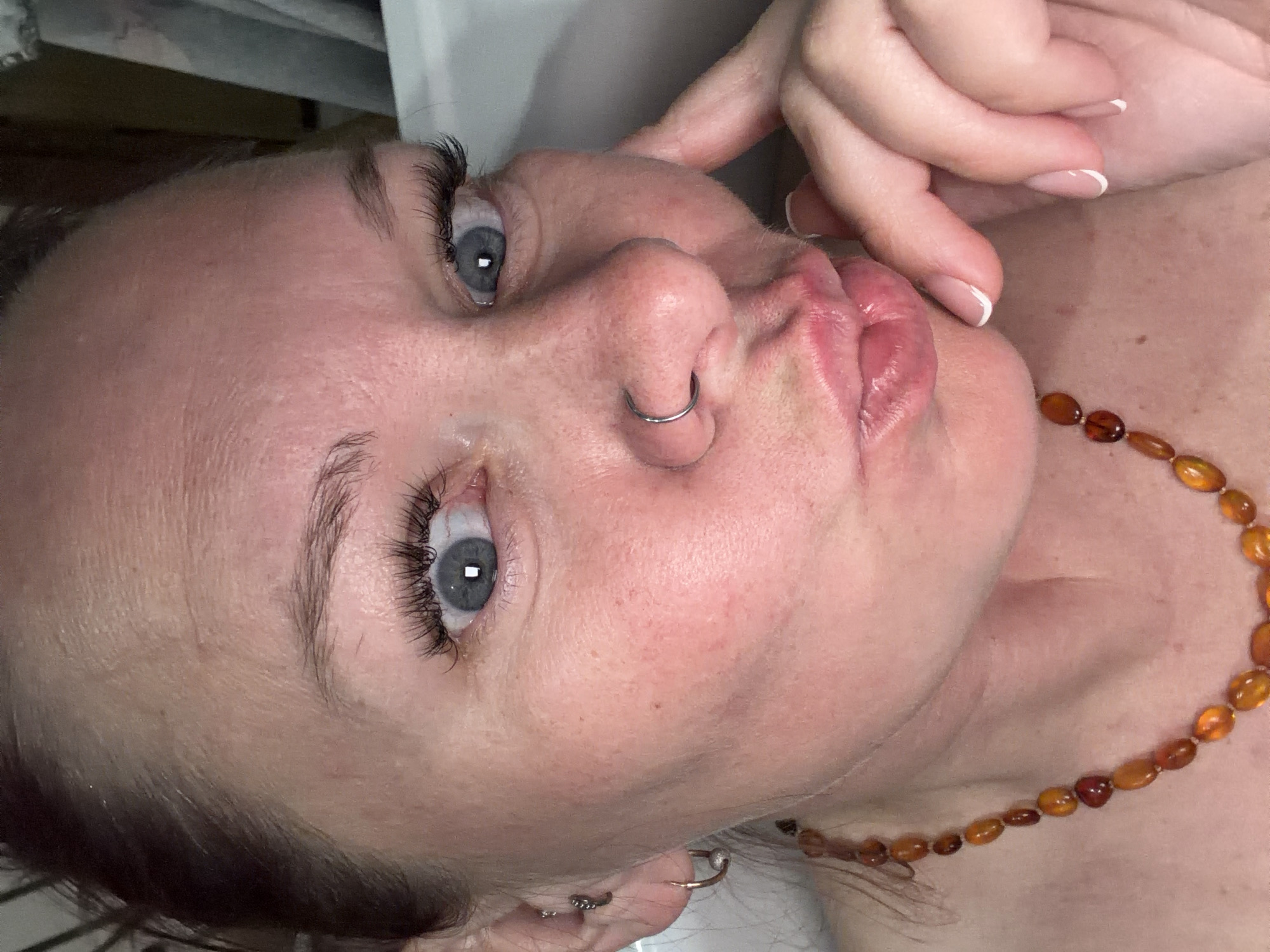 A close-up, angled portrait of a woman with fair skin and light hair. She has blue eyes, a nose ring, and is wearing a necklace of amber-colored beads. Her fingers are near her mouth, and her lips are pursed. Multiple earrings are visible on her left ear. The background is a neutral color. The image does not provide any information pertaining to Chaikovsky, Russia.